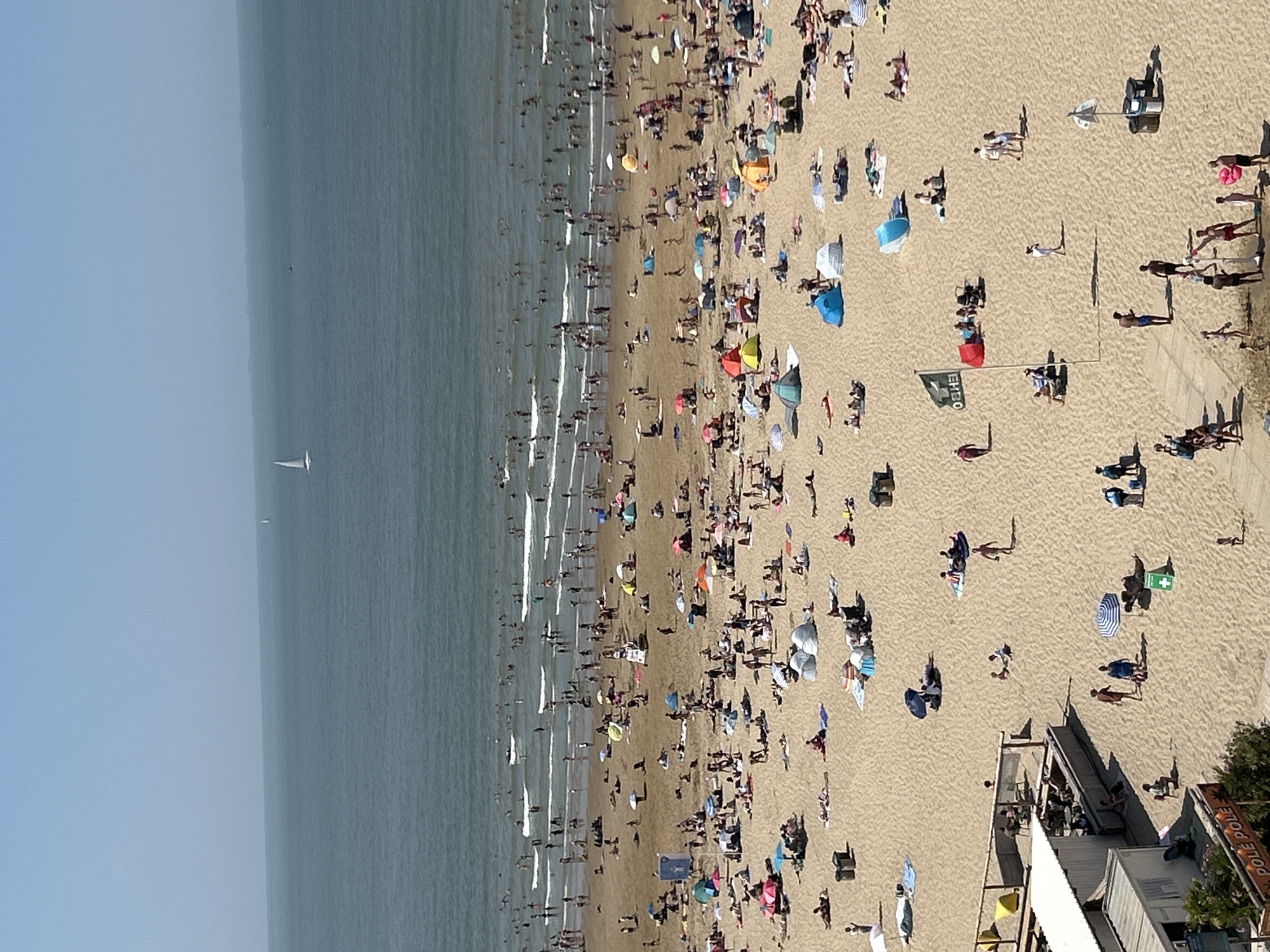 This is an aerial, wide-angle, eye-level view of a crowded beach in Ostend, Belgium. The image captures a large expanse of sand filled with numerous people sunbathing, playing, and relaxing under umbrellas and small tents. The sea is visible in the background, with gentle waves and a few sailboats on the horizon. The sand is a light tan color, and the sky is clear and bright blue.
In the foreground, there are several structures. To the left, a building with a white roof and grey walls, possibly a beach facility or cafe, is partially visible. A sign that reads "POLE POLE" is attached to its side. Further to the right, a section of wooden fencing encloses an area with tables and chairs, where some people are seated. Various beach amenities like trash cans and umbrellas are scattered across the sand.
The beach is densely populated, with individuals and groups spread out across the sand. People are seen in swimwear, with some lying on towels, others walking, and children playing. Colorful umbrellas and beach tents dot the landscape, providing shade for beachgoers. The overall scene depicts a lively and active summer day at the beach.
