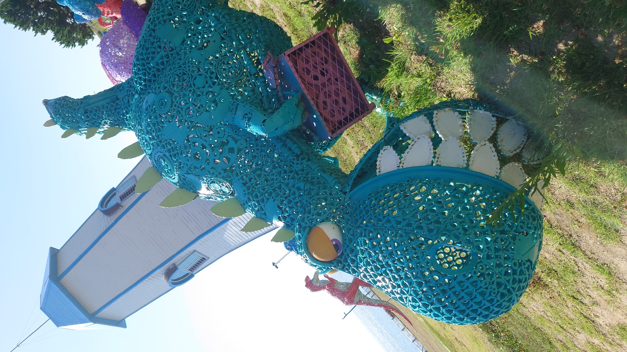 The image captures a vibrant, outdoor scene with a prominent, large-scale turquoise dragon sculpture made of metal. The dragon is detailed, featuring intricate cutouts and spiked elements along its back. In the background, a tall, white, tower-like structure with a blue roof stands at an angle, suggesting it's part of a building or installation. The ground is covered in grass and dirt, indicative of an outdoor park or recreational area.
The sculpture is positioned in a way that it appears to be interacting with the environment. A smaller, pink dinosaur sculpture is visible in the mid-ground, to the left of the turquoise dragon. The sky is clear and bright, suggesting a sunny day.
This particular sculpture is located in the Ulsan Grand Park in Ulsan, South Korea. Ulsan Grand Park is a significant urban park known for its various attractions, including art installations, botanical gardens, and recreational facilities. The dragon sculpture is a notable feature within the park, adding to its whimsical and artistic character. The presence of the tower in the background might be another structure within the park, or possibly a nearby building visible from the park's grounds. The overall atmosphere is cheerful and playful, with the sculptures serving as focal points of artistic expression.