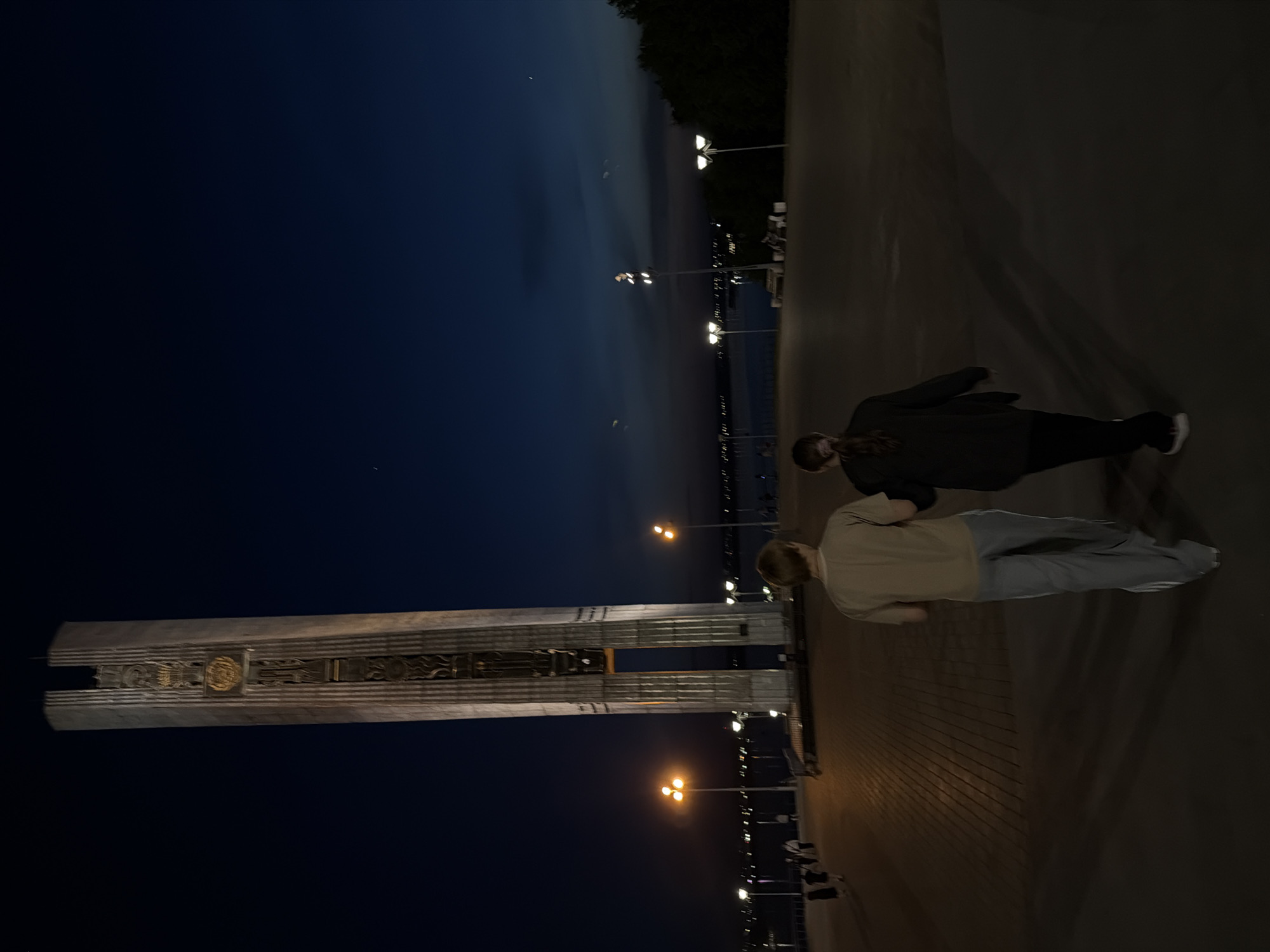 At night in Izhevsk, Russia, two individuals stand on a paved walkway near the "Friendship of Peoples" stele. The stele, tall and featuring decorative insets, is illuminated by nearby streetlights. The individuals, one in dark clothing and one in lighter clothing, appear to be talking. Streetlights line the walkway and extend into the distance along a waterfront. The sky transitions from dusk at the horizon to a dark blue above.
