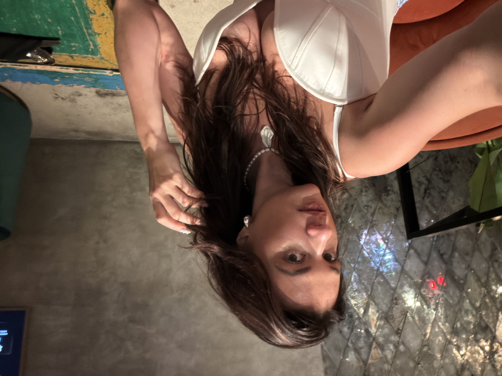 A woman with long, dark brown hair is seated on an orange chair in a dimly lit room. She is wearing a white corset-style top and a pearl necklace with a silver pendant. Her right arm is raised, and her hand is touching her hair. The background features a textured wall with geometric patterns and a concrete floor with patches of blue, yellow, and green paint. A dark-colored object, possibly a bag, is visible on the floor. The scene appears to be indoors, potentially in a restaurant or lounge setting.
