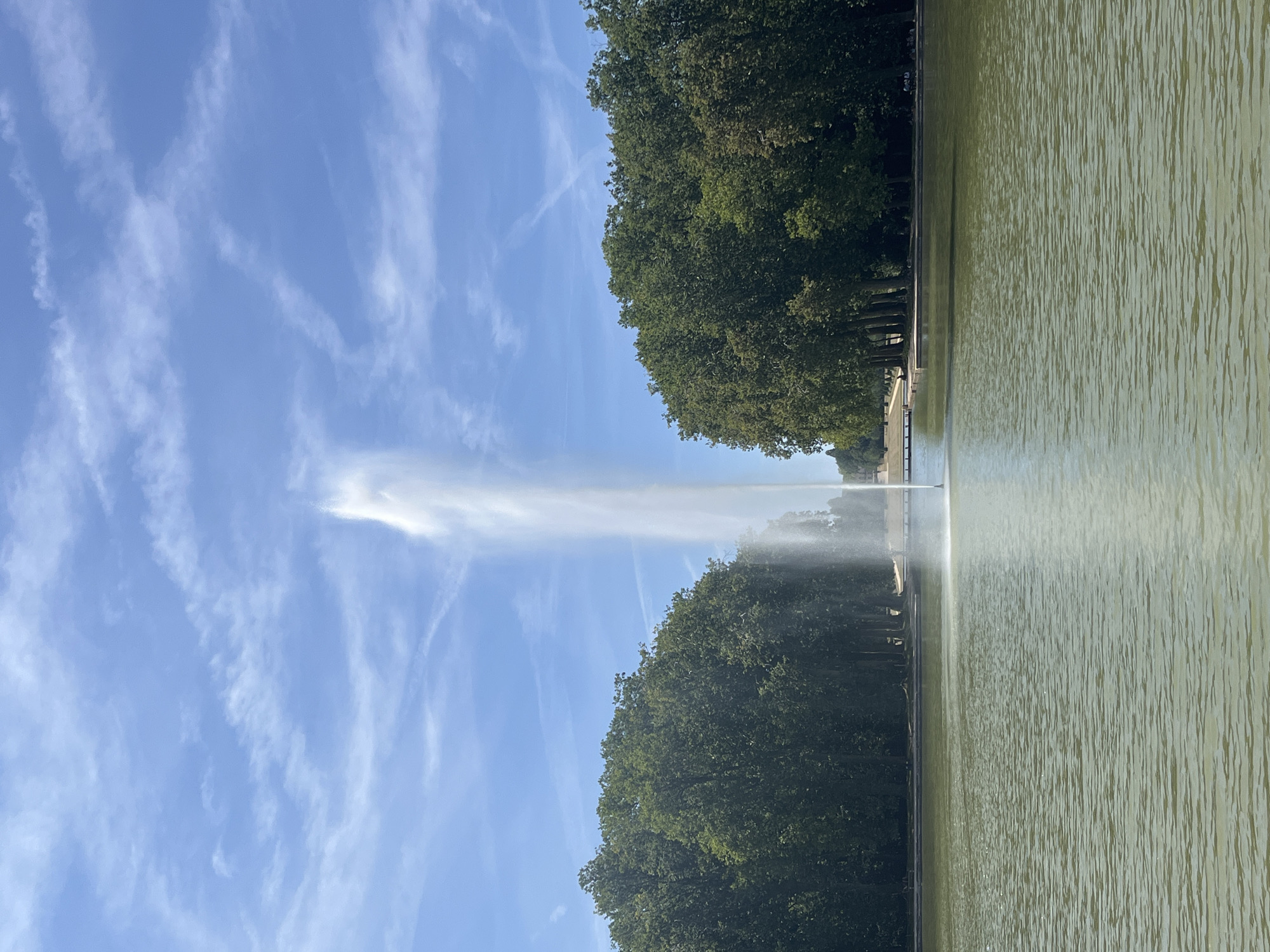 The image depicts a large, vertical water fountain erupting from the center of a wide, calm body of water. The water in the pond is a greenish hue, with subtle ripples on its surface. On either side of the pond, dense green trees form a natural border, their foliage thick and lush. Beyond the trees, a clear blue sky with thin, wispy white clouds stretches across the frame. The fountain's spray reaches high into the sky, creating a dramatic vertical element against the backdrop of the trees and sky. In the distance, a long, paved walkway or embankment is visible, with a few small figures and structures suggesting a park or garden setting. This scene is located at the Parc des Sceaux in Antony, France.
