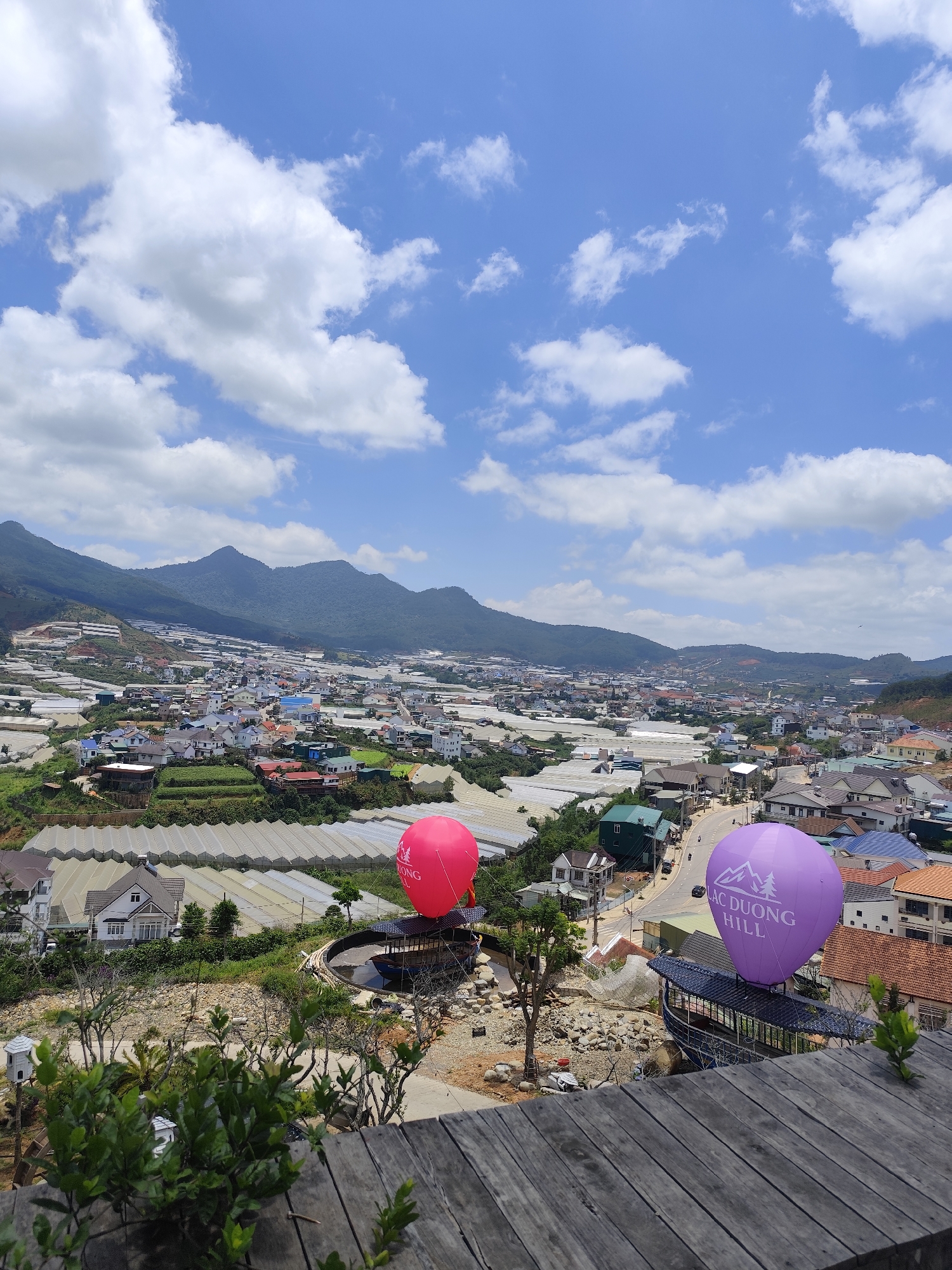 The image depicts a landscape scene in Lạc Dương, Vietnam. Mountains form a backdrop to a town with numerous houses and greenhouse-like structures. In the foreground, there is a wooden deck or viewing platform. A pink and a purple balloon, respectively, are positioned on platforms. The purple balloon has the words "LAC DUONG HILL" printed on it. A road winds through the town, with houses and greenhouses lining the sides. The sky is blue with scattered white clouds.