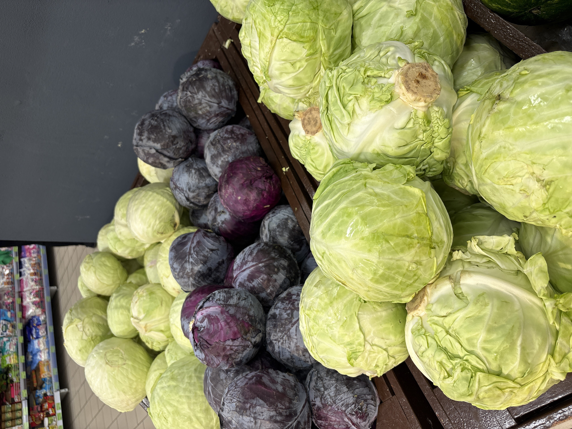 The image shows a display of cabbages, both green and red, in a store. The cabbages are piled in a dark wood crate. In the background, there are shelves with various items, suggesting that this photo was taken inside a shop. The exact location in Minsk, Belarus is not explicitly stated or visually identifiable from the given image.