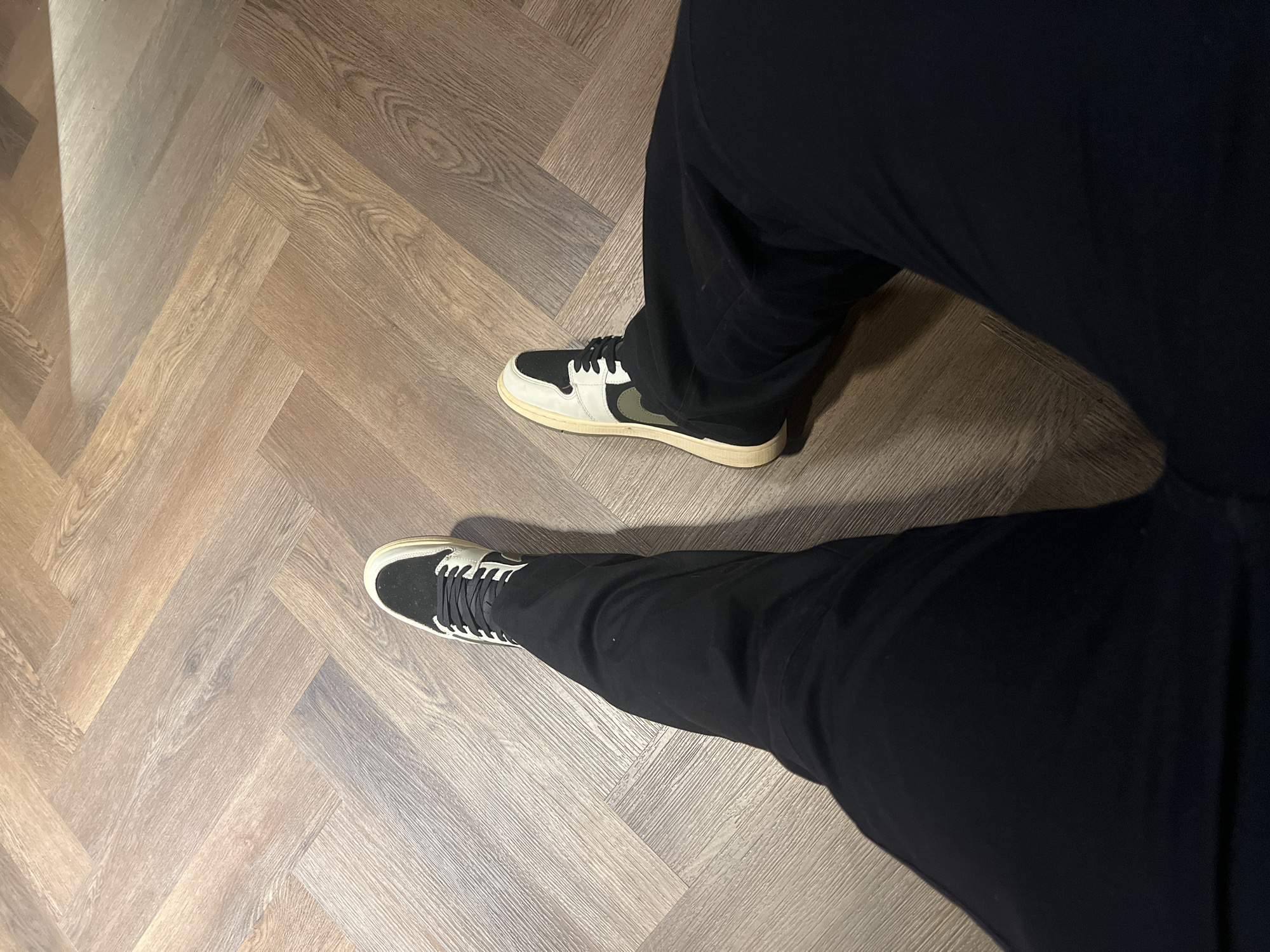 The image captures a top-down view of a person's lower legs and feet, set indoors in Abuja, Nigeria. The individual is wearing black trousers that extend down to the ankles. Two distinct sneakers are visible on a light brown wooden floor, arranged in a herringbone pattern.
The left sneaker (from the viewer's perspective) features a predominantly black upper with white laces and a white trim outlining the toe box and side panels, all resting on a cream-colored sole. The right sneaker has an off-white main body, a black toe cap, black laces, and a prominent dark olive green swoosh-like emblem on its side, also set on a cream-colored sole.
The person appears to be standing or in the process of walking. The indoor lighting casts soft shadows around the legs and shoes on the textured wooden floor. There is a slightly brighter area on the floor in the upper left portion of the image. No specific time of day or weather conditions are discernible, given the indoor setting. No visible text is present within the frame.