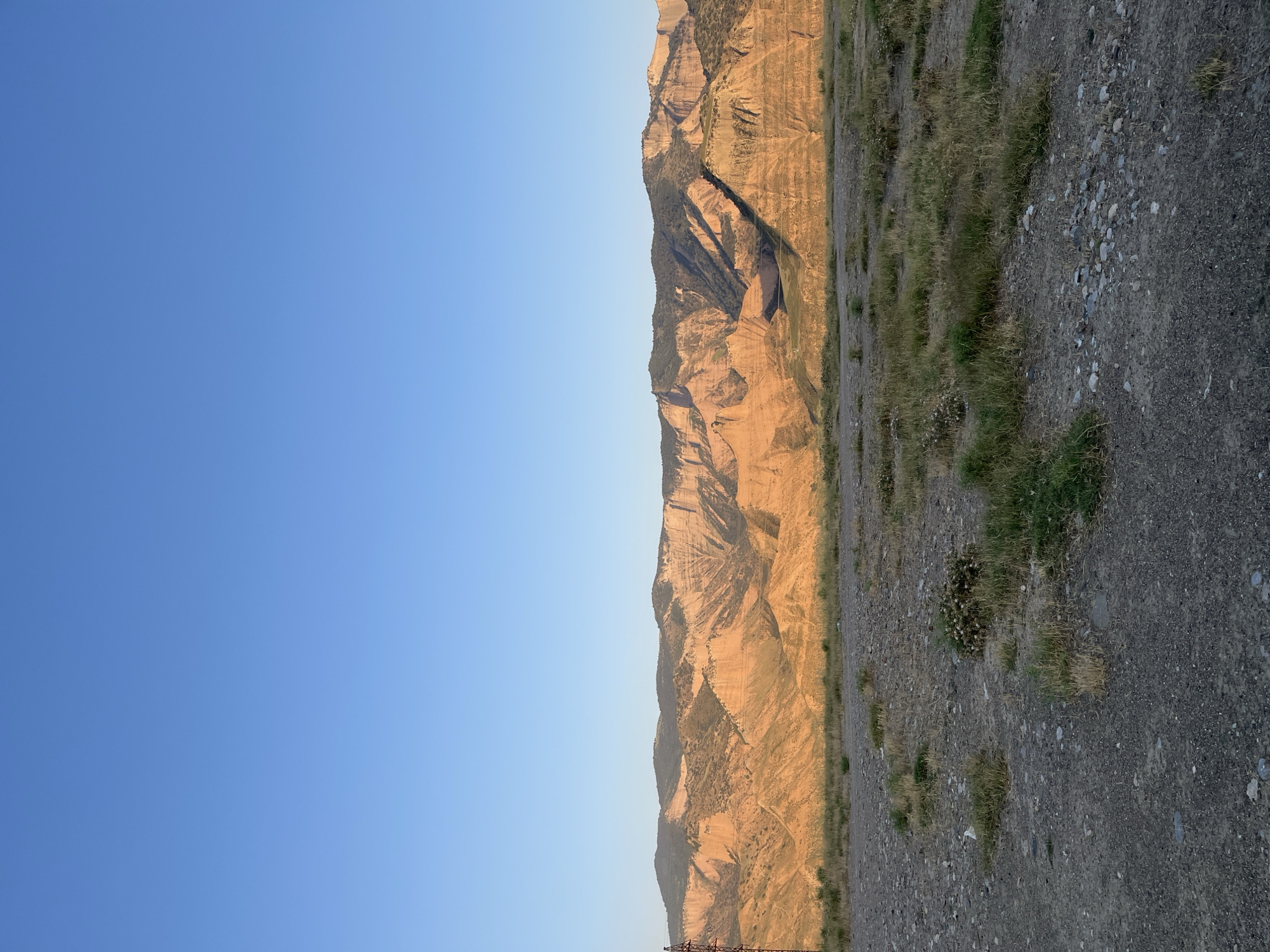 The image displays a wide, arid landscape under a clear blue sky. In the foreground, a rough, gravelly ground is sparsely covered with dry grass and some low-lying vegetation. A metallic, lattice-like structure, possibly a utility pole or tower, stands tall on the left side of the frame.
Dominating the background is a vast mountain range, characterized by layered rock formations exhibiting shades of orange, brown, and gray. The peaks are sculpted by erosion, creating a rugged and textured appearance. The setting sun casts a warm, golden light across the mountains, highlighting their geological features. The overall scene suggests an undeveloped, remote area with a focus on natural terrain. The image is devoid of any people or overt signs of urban activity, other than the aforementioned structure.