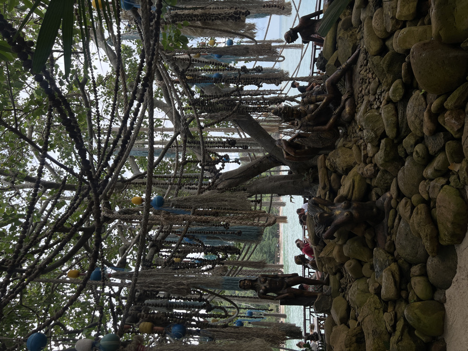 This image depicts an outdoor seaside establishment in Baan Sri Thanu, Thailand. The foreground consists of a sandy patch leading to an arrangement of large, dark, irregular rocks that form a naturalistic base. Integrated into these rocks are five bronze-toned statues: a standing nude female figure on the left, a kneeling nude female figure with raised arms in the lower center, two intertwined figures adorned with elaborate headwear seated together in the right-center, and a partially visible reclining figure on the far right.
Dominating the scene is a large, multi-trunked tree with extensive aerial roots or branches heavily decorated. Hanging from the tree are numerous strands of material including natural fiber ropes, bundles of light-colored fibrous material resembling dried grass or nets, and long strings of dark spherical beads. Interspersed among these are spherical floats or ornaments in shades of blue, yellow, and light green.
In the midground, behind the statues and rocks, is a dark-colored wooden deck or platform situated at the edge of a calm body of water. Several small tables and chairs are visible on this deck. Approximately six to eight individuals are seated at these tables, appearing to be dining or socializing.
The background reveals a tranquil bay with light blue-green water, a narrow strip of sandy beach, and lush green hills covered in vegetation. The sky, visible through the tree canopy, is overcast and pale. The scene is illuminated by diffused daylight, casting soft shadows.