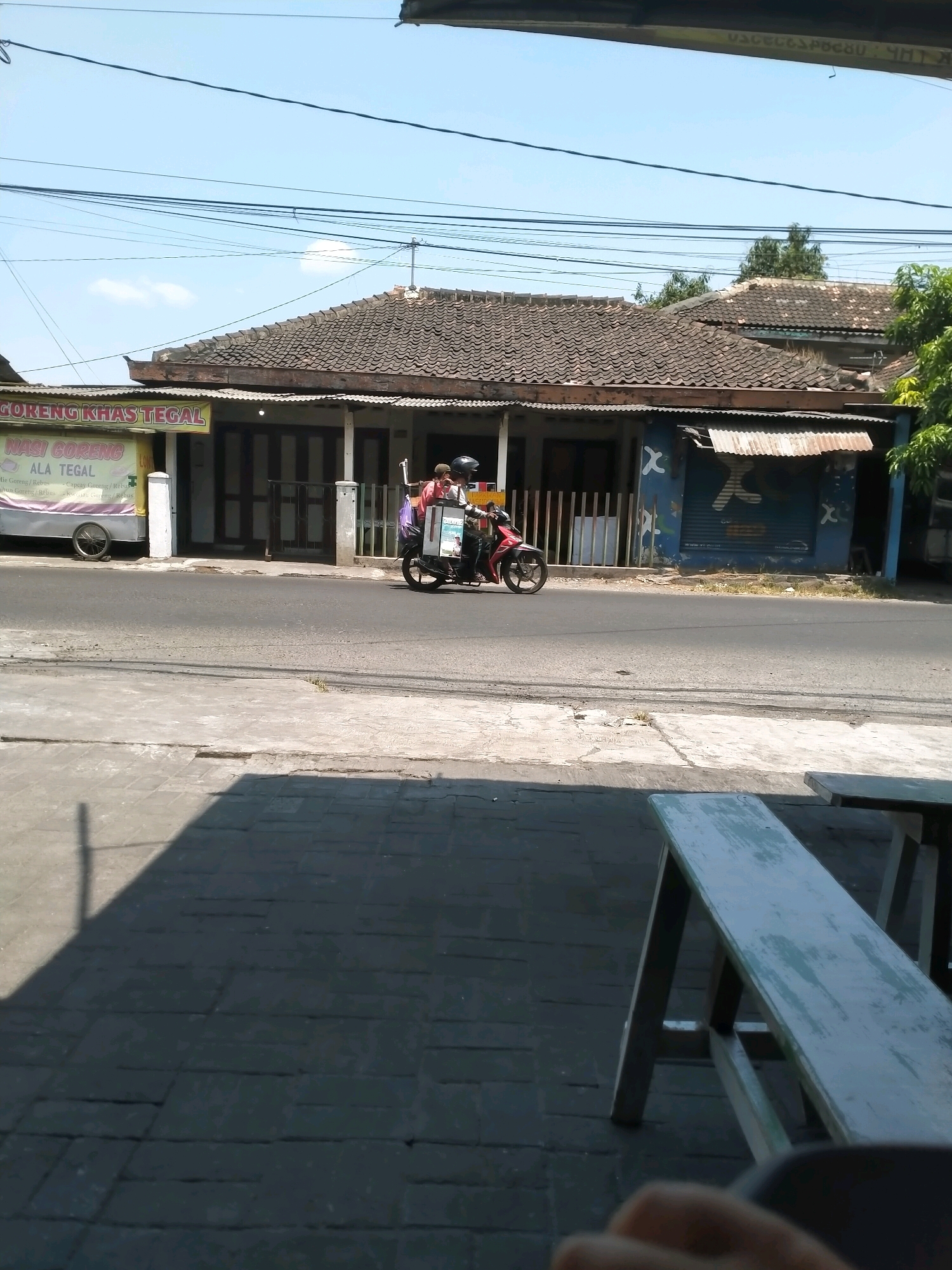 The image captures a street scene in Kasihan, Indonesia, on a clear, sunny day. In the foreground, a shaded area shows a dark grey brick-paved walkway and a weathered, light blue wooden bench. The middle ground features a paved road with a red motorcycle carrying two people driving from right to left. The rider wears a helmet, and a passenger behind them also wears headwear. The motorcycle is adorned with a white cooler box on the rear. Adjacent to the road is a sidewalk with a white pillar. Behind the sidewalk, a building with a tiled roof has an awning covering a storefront. A food cart with a yellow sign is parked to the left of the building. The building's facade includes dark doors and a blue section with white 'X' symbols. A streetlamp is visible on the building's right side. Power lines are strung across the sky above the building. Trees are present on the far right. The background displays a blue sky with a few scattered white clouds.
