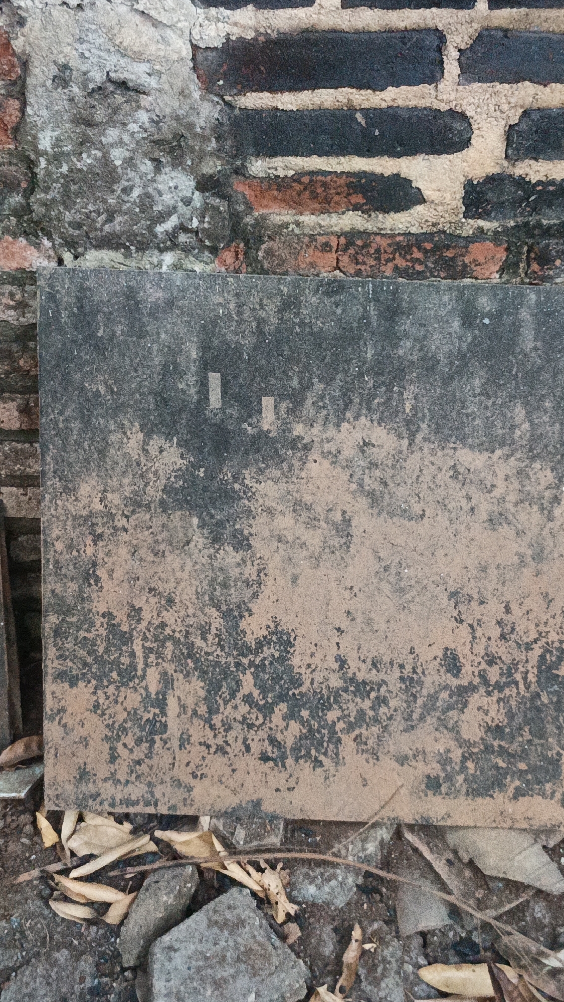 The image shows a close-up view of a weathered wall and a concrete slab in Kandangmas, Indonesia. The wall is constructed of bricks, some of which are dark and appear aged, interspersed with lighter-colored mortar. A section of the wall to the left has a rough, textured surface with patches of gray and white. Below the brick wall, a large, flat concrete slab rests on the ground. This slab is dark gray and covered in a mottled pattern of lighter, sandy-colored material and dark speckles, indicating wear and dirt accumulation. Two small, rectangular, lighter-colored markings are visible on the upper portion of the slab. The ground in front of the slab is covered with loose debris, including gray stones, dry brown leaves, and twigs. The scene is lit from above, casting subtle shadows on the textured surfaces. There are no people or animals visible in the image.