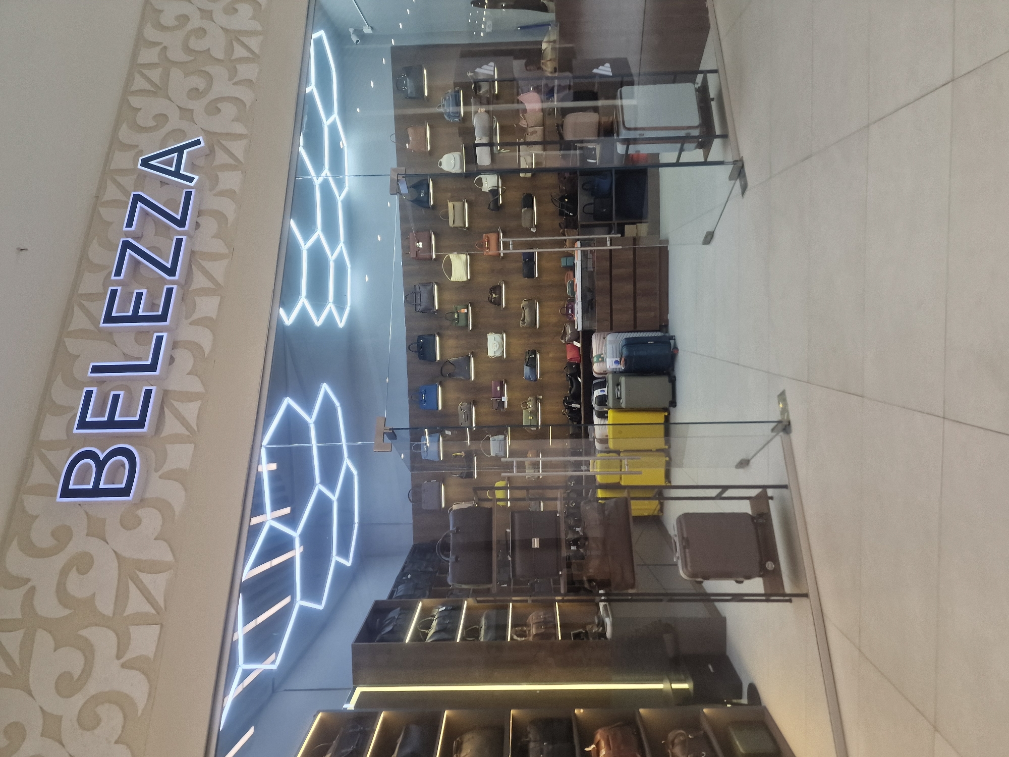 The image shows the exterior of a store called "BELEZZA" located in the city of Kokshetau, Kazakhstan. The store entrance features large glass doors and windows, offering a view of the interior. Inside, the store is filled with a wide variety of handbags and luggage displayed on shelves and racks. The store's signage, "BELEZZA", is prominently displayed in large, illuminated letters above the entrance. The interior lighting is modern, with hexagonal LED fixtures illuminating the display areas. The store appears to specialize in fashion accessories, particularly bags and travel suitcases, with a selection of brightly colored suitcases visible near the entrance. The floor is tiled, and the overall aesthetic is contemporary and clean.