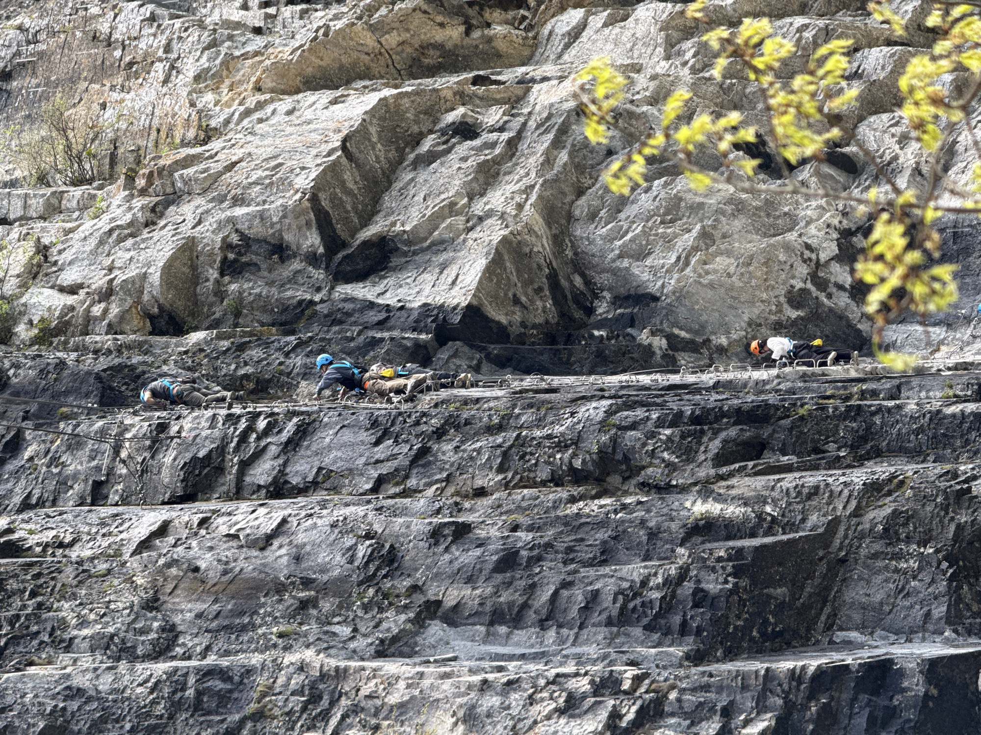 The image shows a group of people on a via ferrata route on a steep rock face. The location is the city of Yongding District, China. The people are wearing climbing helmets and harnesses and are attached to a steel cable that runs along the rock face. The rock face is made up of dark gray stone and is characterized by horizontal ledges. The individuals are actively engaged in traversing the via ferrata, navigating along the metal rungs and cables. The environment appears to be a quarry setting. Vegetation is visible in the background.
