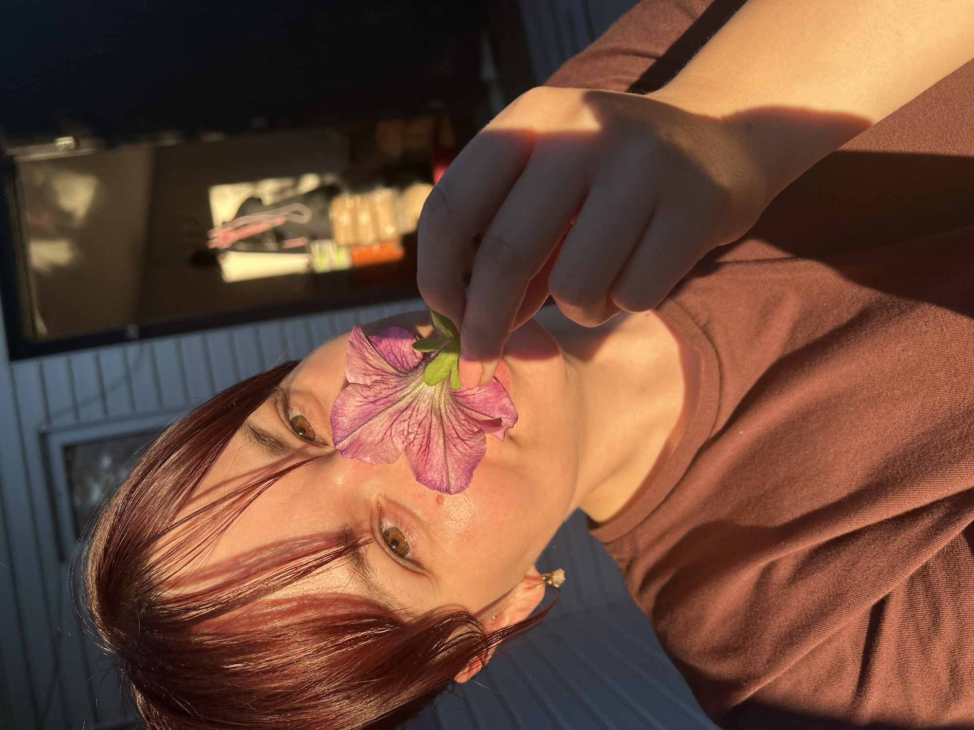 This media file shows a close-up of a person's face and hand, with a purple flower held near their mouth. The person has reddish-brown hair and brown eyes. The lighting suggests it is late afternoon or early evening, with warm sunlight casting shadows. The background is blurred but appears to be outdoors, with some structures visible. There is no visible text in the image. The overall mood is intimate and serene.