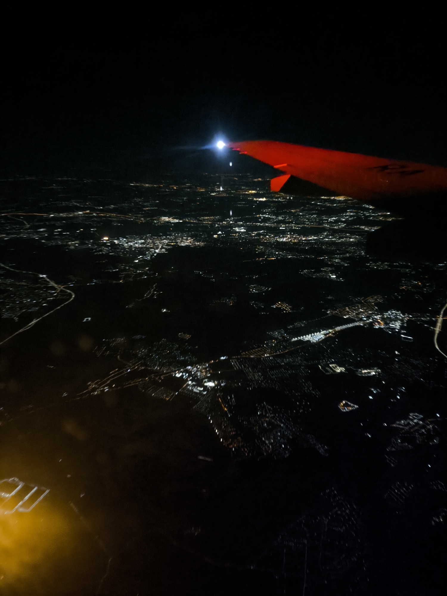 The image shows a night view from an airplane, with the wing visible in the upper right corner. The wing is red and has a bright white light on its leading edge. Below, the city lights of Khimki, Russia, are visible as a scattered network of bright spots and streaks. The lower left corner of the image is obscured by a blurry yellow light, possibly from inside the airplane. The sky above is completely dark.