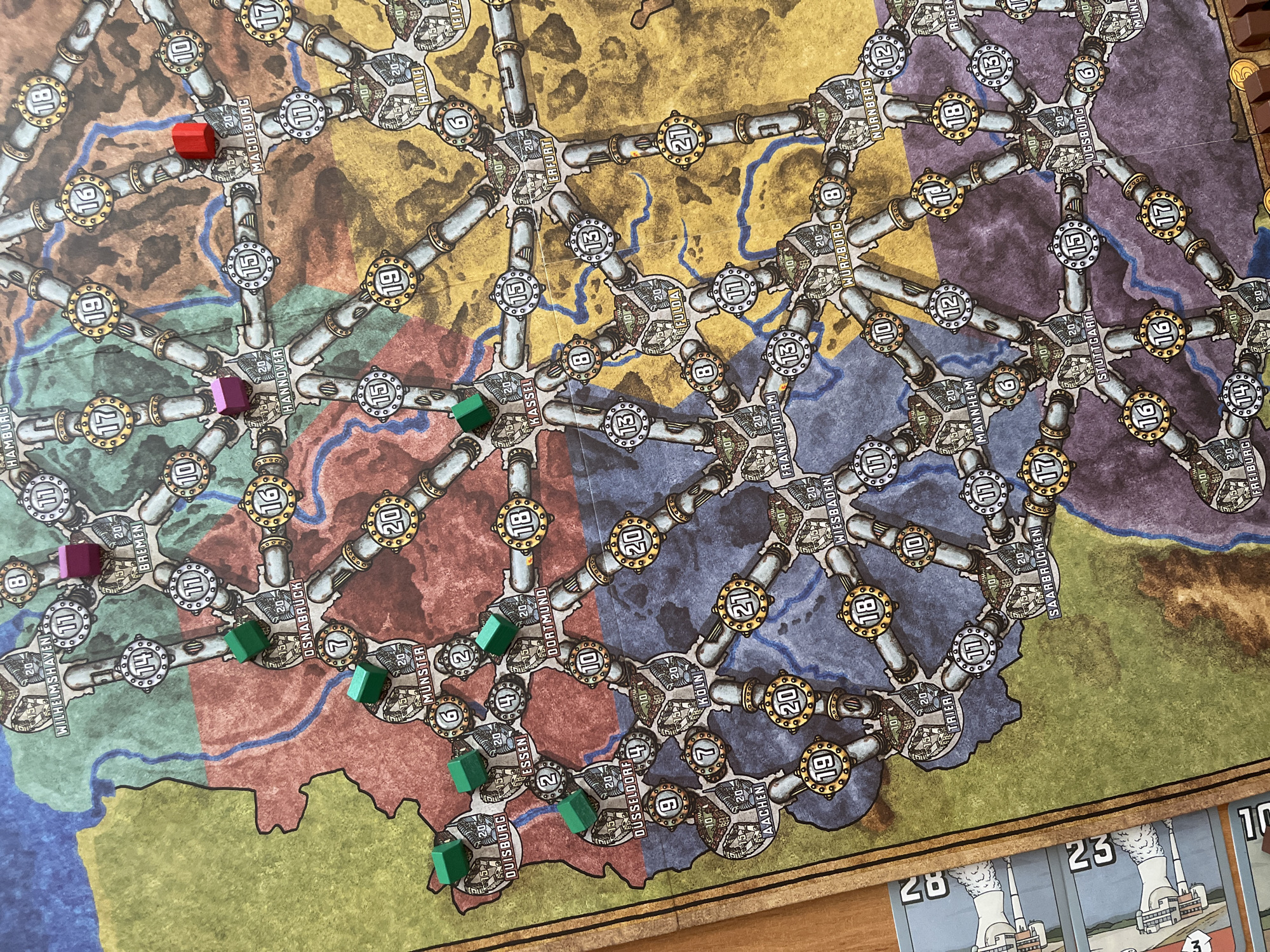 The image shows a board game with a map of Germany, divided into color-coded regions. Several wooden game pieces in green, red, and purple are placed on the map, marking different cities and routes. The cities are labeled with names like "Wilhelmshaven", "Bremen", "Hannover", "Magdeburg", "Duisburg", "Essen", "Dortmund", "Münster", "Köln", "Frankfurt", "Stuttgart", and "Munich". The game board appears to be a network of train routes connecting these cities. There are no elements in the image that relate to the city of Taraz, Kazakhstan, or any form of city administration there.
