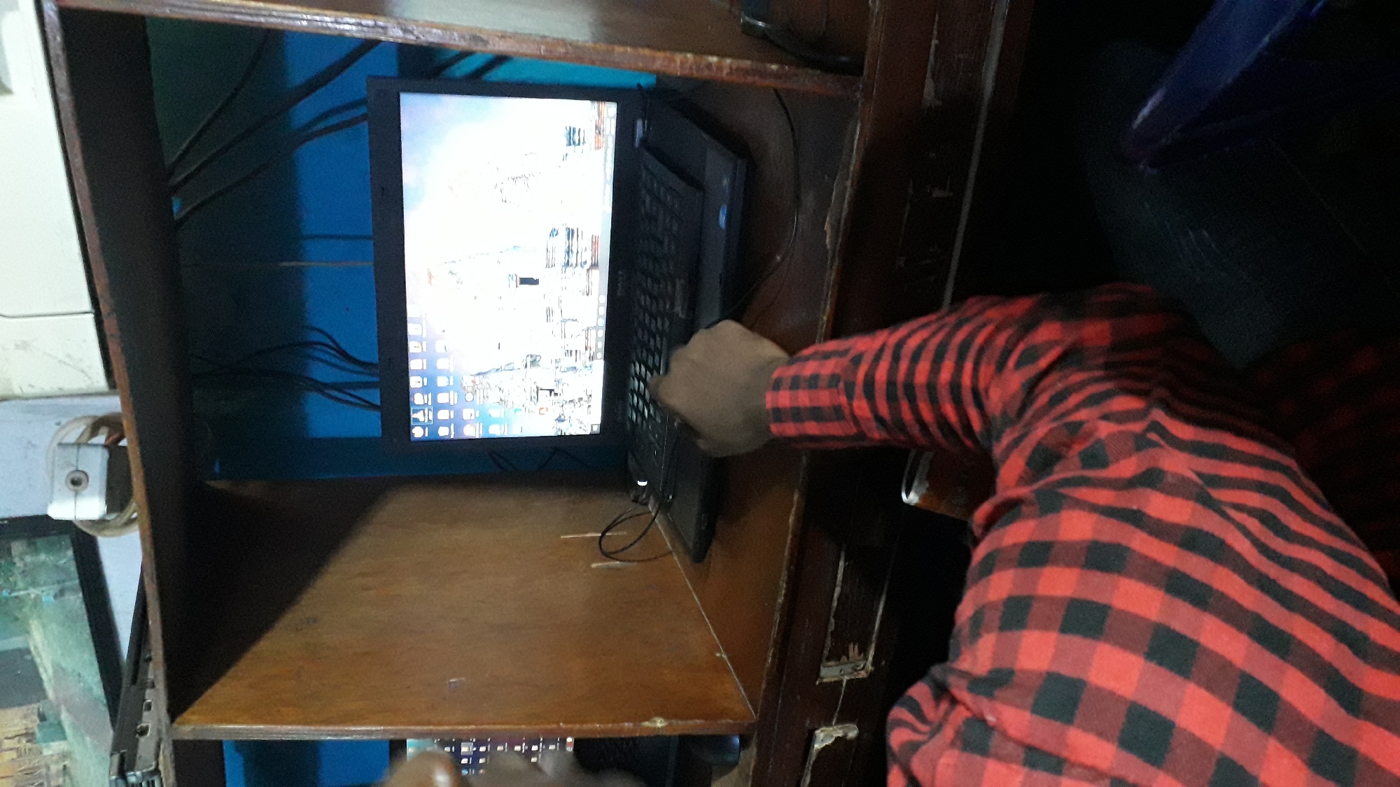 In Onitsha, Nigeria, a person wearing a red and black checkered shirt is using a laptop at a wooden desk. The laptop is open, displaying a desktop background with what appears to be a digital image of a city or architectural structure. Wires are visible behind the laptop, connected to the computer and possibly other devices. The desk is part of a cubicle-like setup, suggesting an internet cafe or similar workspace. The lighting is dim, with the laptop screen being the primary light source in the immediate foreground. The person's hand is on the keyboard, indicating active use of the computer.