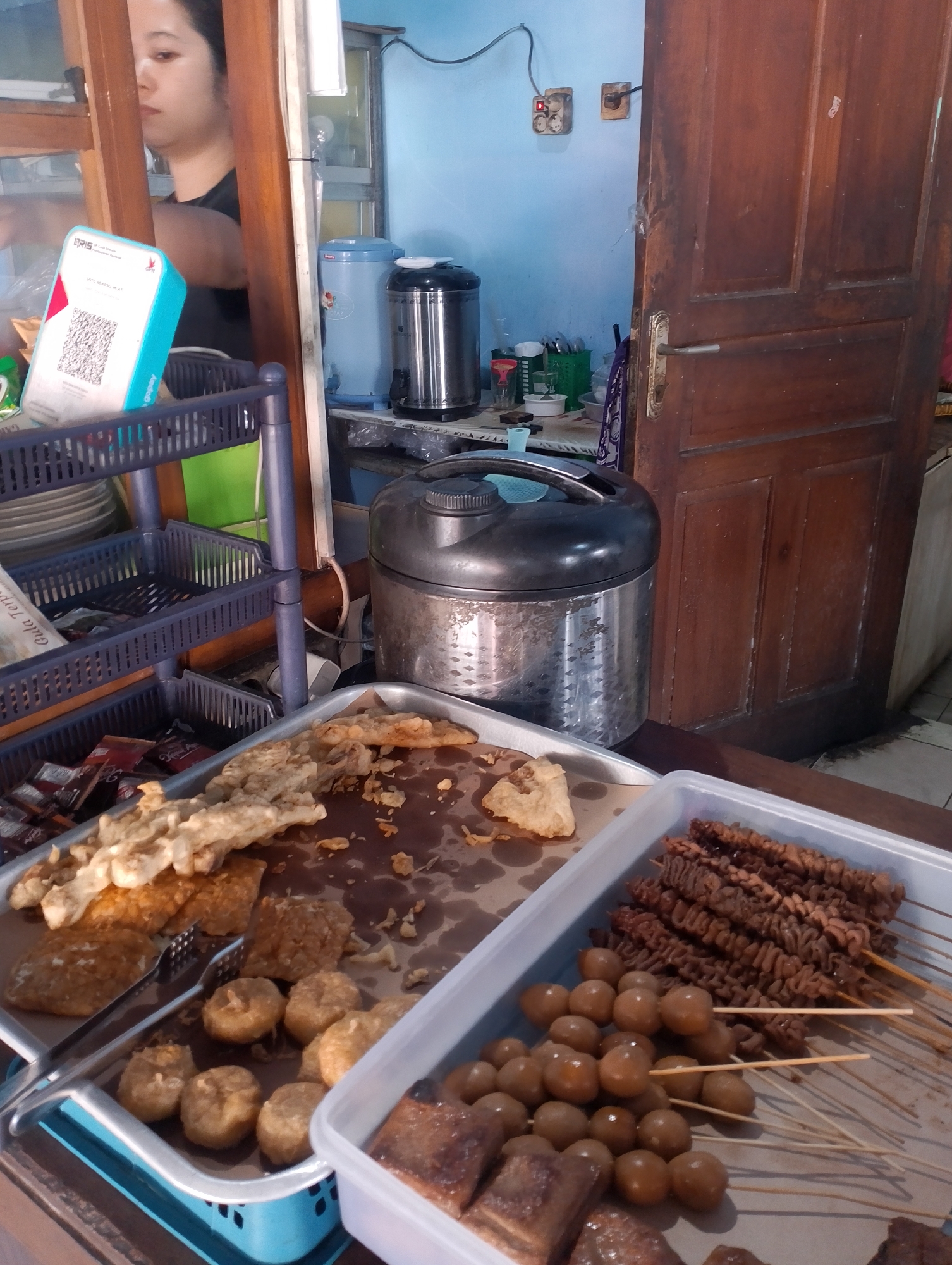 The image depicts a food stall in Sinduadi, Indonesia. In the foreground, a selection of fried foods is displayed in plastic containers and on a metal tray. These include what appear to be fried tempeh and some round, bite-sized fried items. A separate container holds skewers of what looks like grilled meat intestines and quail eggs. A pair of metal tongs rests on the tray of fried items. Behind the food, a woman is partially visible through a wooden frame, appearing to be preparing or serving food. Several plastic baskets holding various items, including what looks like packaged snacks and possibly plates, are stacked to the left. In the background, a large stainless steel thermos and a blue water dispenser are visible next to a wooden door. Electrical outlets and switches are mounted on a light blue wall. The overall scene suggests a busy street food environment.