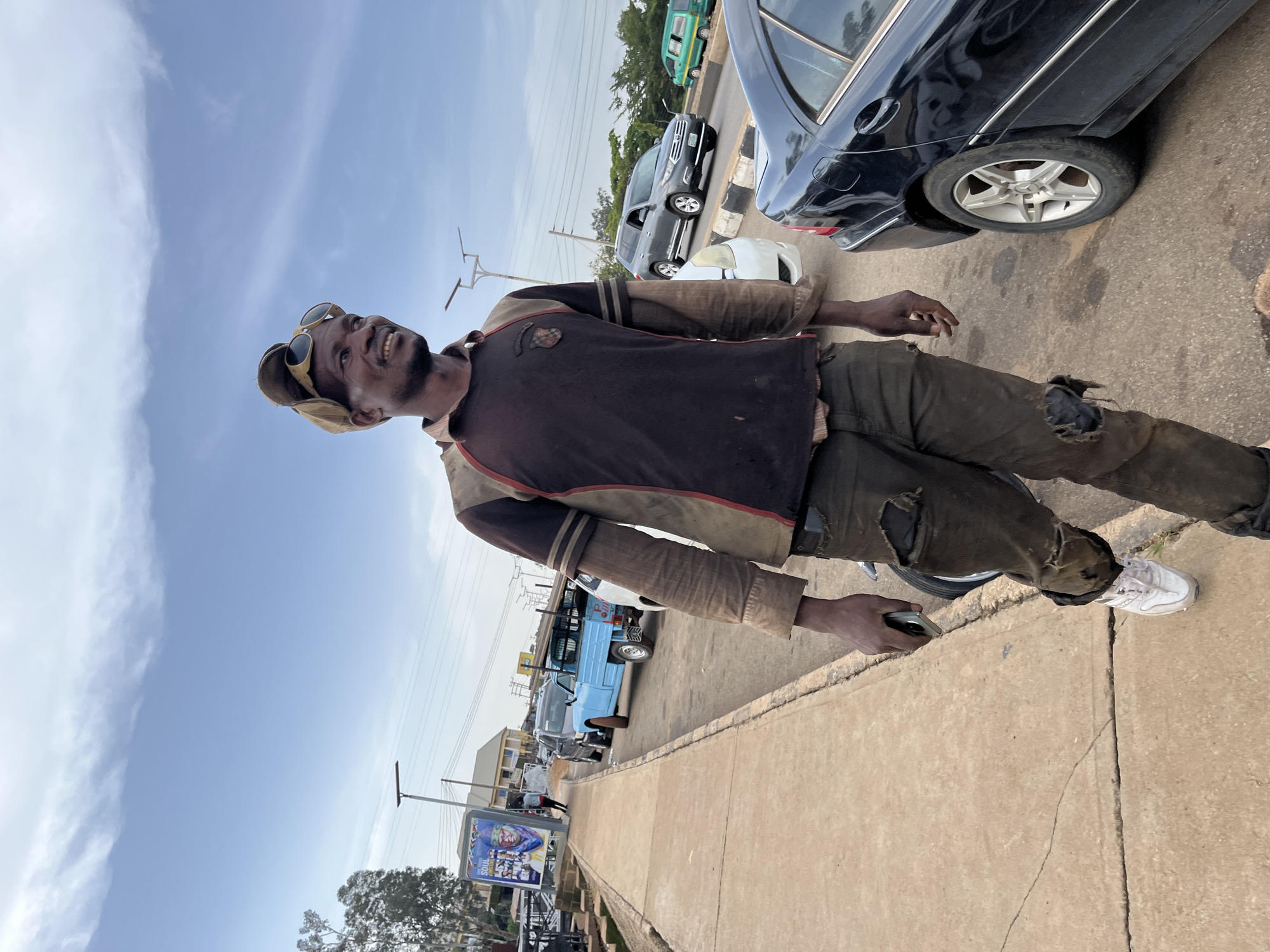 A man stands on a sidewalk next to a street with several cars. He is wearing a brown and maroon jacket, ripped brown pants, and white sneakers. He has sunglasses pushed up on his forehead and is looking upwards with a slight smile. A black car is parked on the street next to him, and other cars and a blue truck are visible further down the road. Buildings and trees can be seen in the background. A billboard with an advertisement is also visible on the left side of the image. The sky is blue with some scattered clouds, suggesting it is daytime. The scene appears to be in a town or city setting, possibly in Gura Top, Nigeria, given the context. The man's posture and expression suggest he might be waiting or observing something.