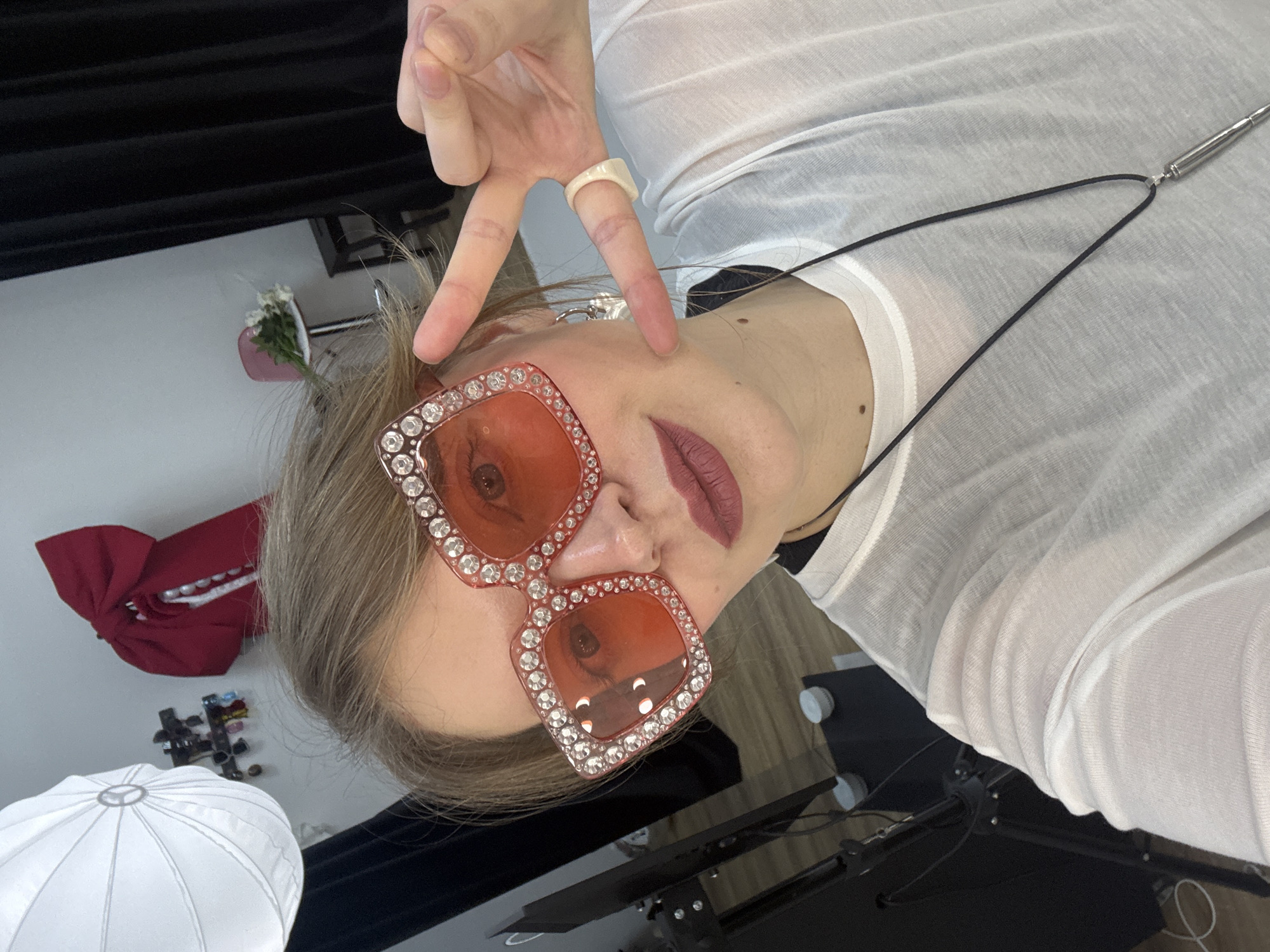 A person wearing large, pink, jeweled sunglasses and a sheer white shirt is making a peace sign with their hand. The person has light-colored hair and is wearing a white ring. They also have a necklace with a cylindrical pendant. Behind the person, there is a black background, some studio lighting equipment, a red bow with pearl accents on a wall, and what appears to be a small bouquet of flowers in a pink container. The location is not discernible from the image alone. There is no evidence to suggest the photo was taken in Izhevsk, Russia.