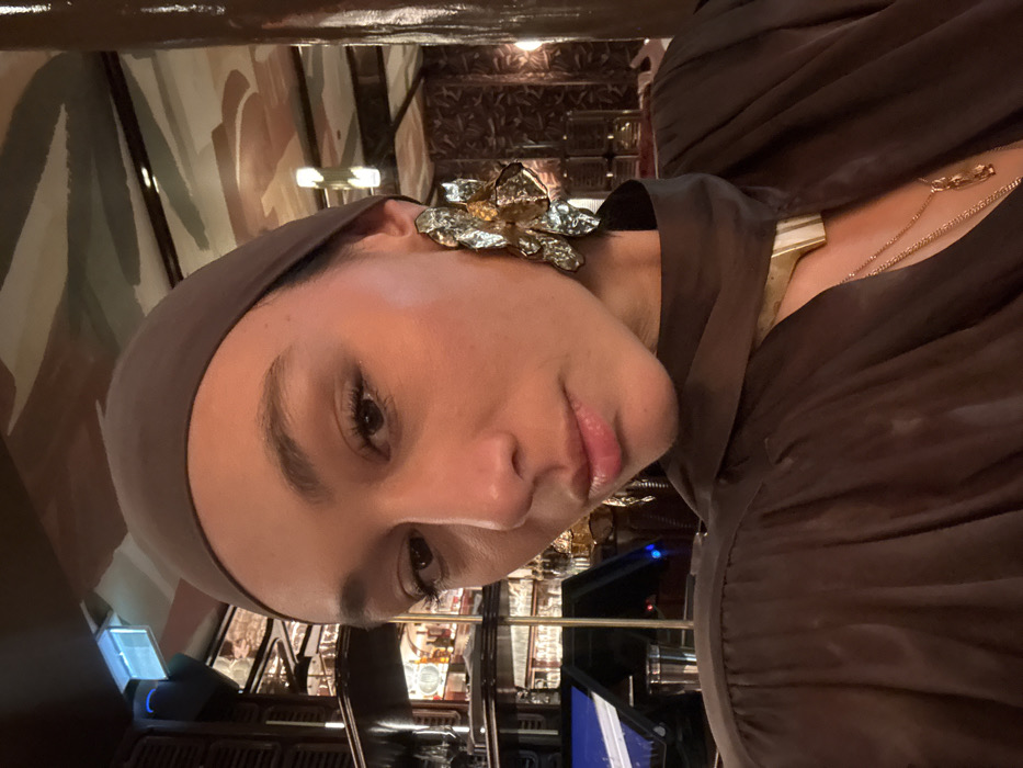 A woman with brown hair and a matching headscarf poses for a selfie. She wears a large silver flower earring, a gold and white collar necklace, and a delicate gold chain necklace. She has a smooth complexion and pink lips. The background reveals a dimly lit restaurant or bar setting, with shelves of bottles and bar equipment visible. The lighting is warm and ambient, contributing to the overall intimate setting.