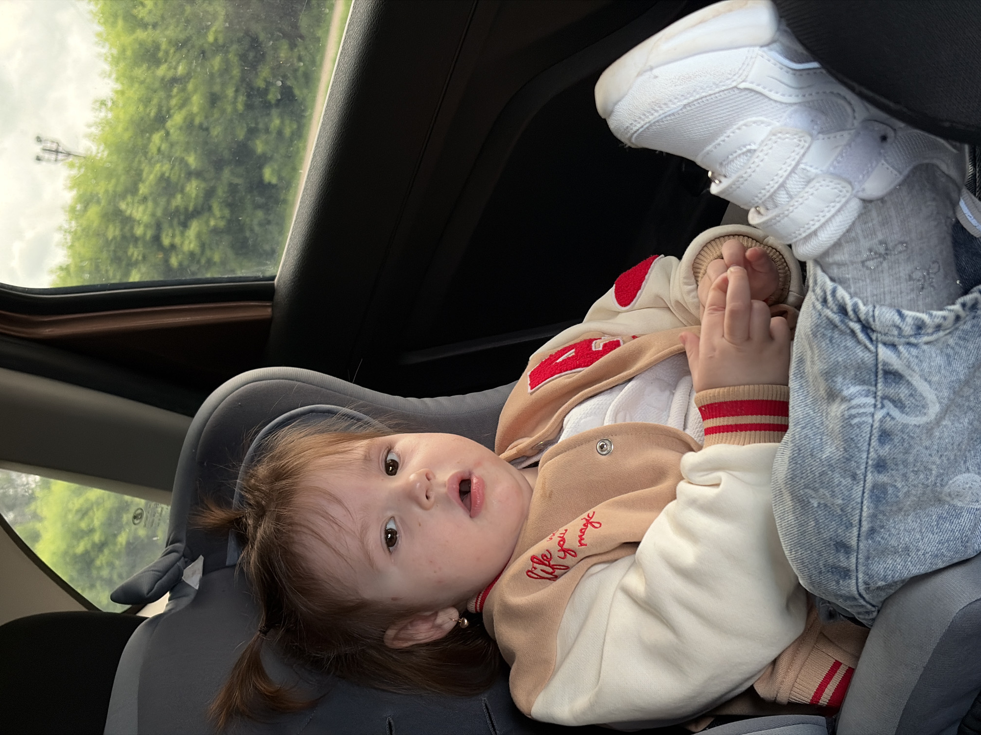 In a vehicle, a young child with auburn hair tied into two pigtails sits in a car seat. The child wears a tan and cream-colored jacket with red trim and lettering, blue jeans, and white sneakers. The child's left foot is propped up on the car seat. Through the car window, a lush green forest and a cloudy sky are visible. A distant power line pole can be seen against the sky. The interior of the car, including the dark-colored door panel and the gray car seat, is also visible. The scene appears to be located in the city of Chaikovsky, Russia, given the context of the image.