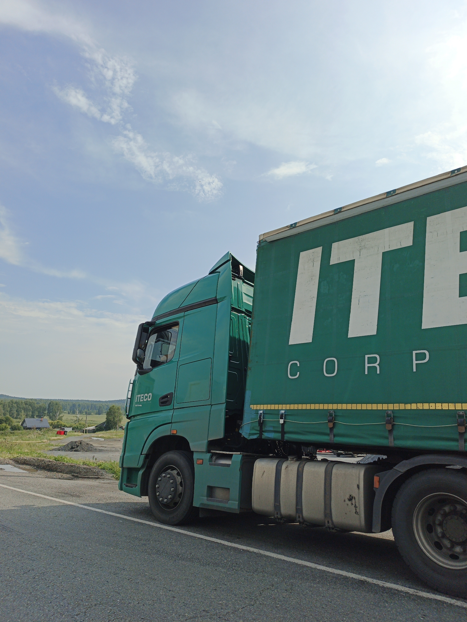 A green semi-truck is parked on the side of a road. The truck has a large green trailer with the white letters "ITECO CORP" printed on the side. The truck cab is also green and has "ITECO" printed on the door. The truck is facing to the right of the frame, and the trailer is extending out of the right side of the frame. In the background, there is a field with some trees and a few buildings. The sky is blue with some white clouds. The location is Nizhnii Atlyan, Russia.