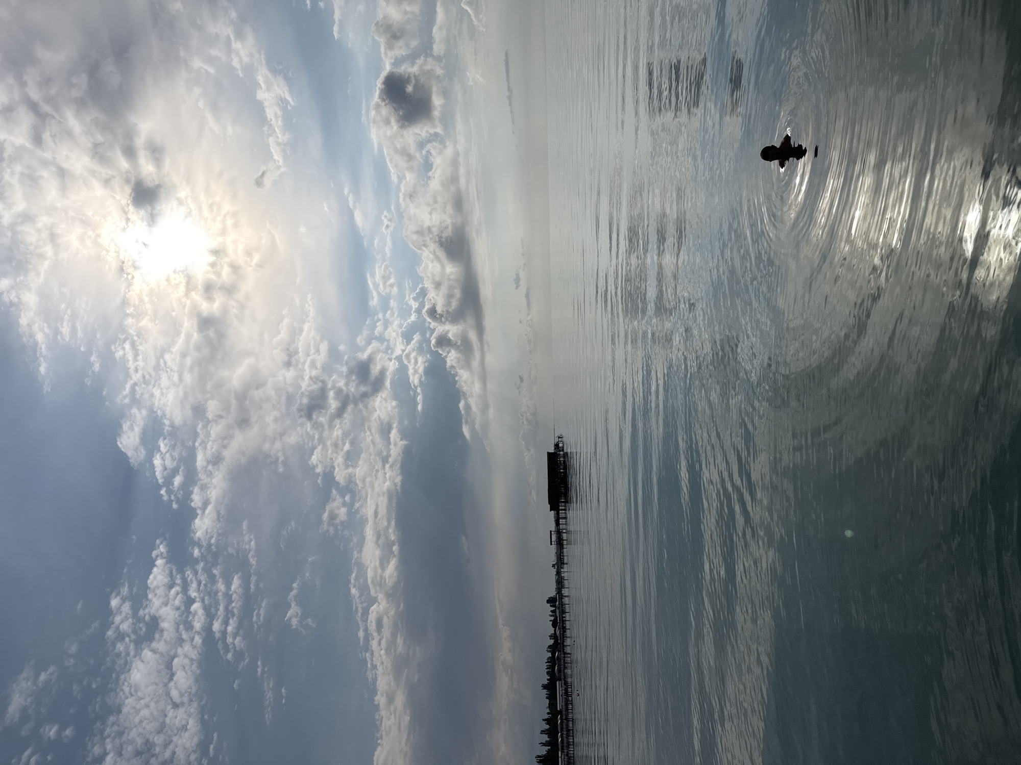 A man swims in the calm, rippling waters of Issyk-Kul lake, near the shore of Sary-Oy, Kyrgyzstan. The sun, partially obscured by scattered clouds, casts a bright reflection on the water's surface, creating shimmering patterns. In the distance, a long wooden pier stretches out into the lake, lined with a row of buildings or cabanas, and a larger structure stands at the end of the pier. The sky is a mix of blue and white, with dramatic cloud formations adding texture and depth. The overall scene evokes a sense of tranquility and natural beauty.