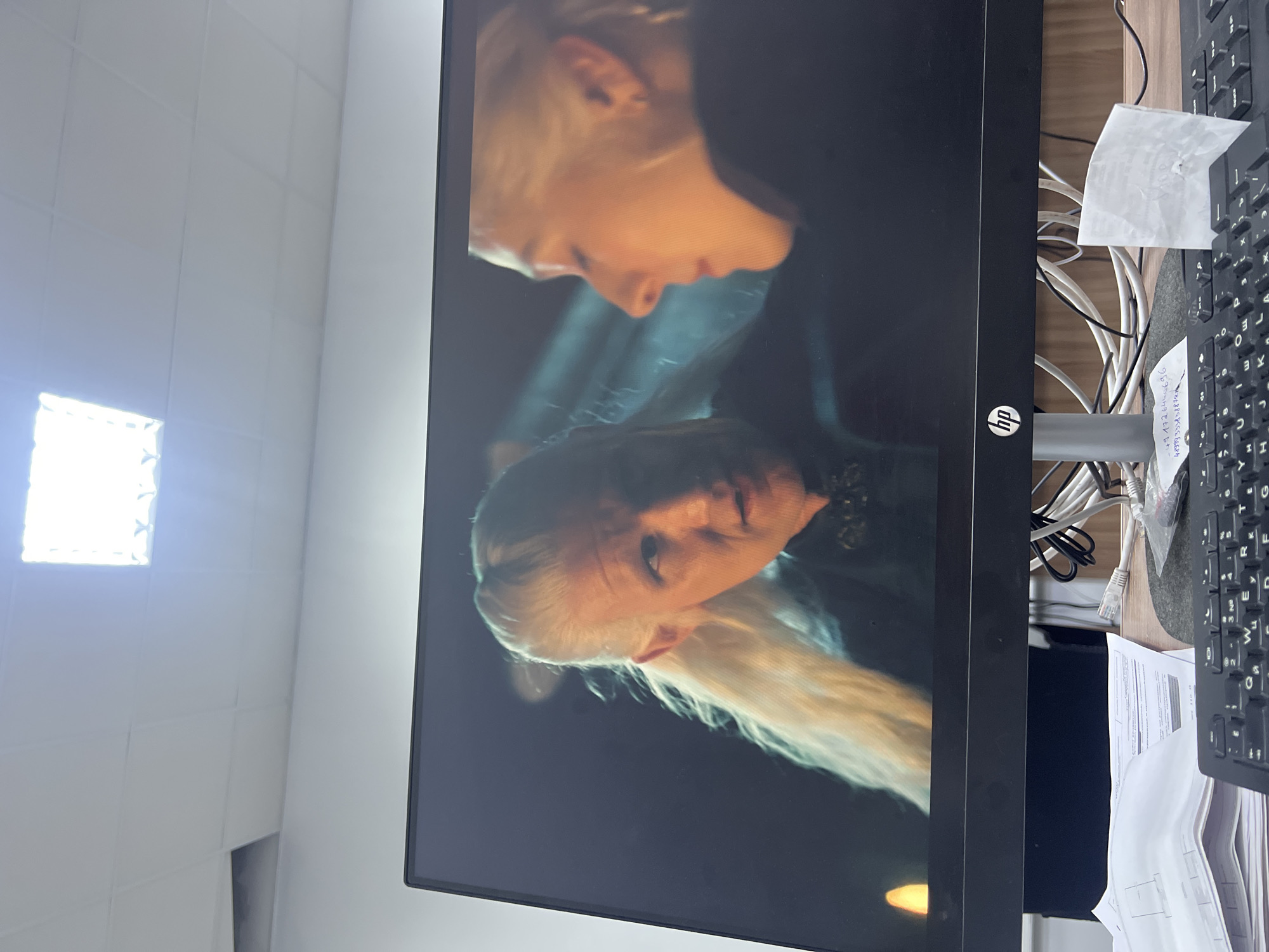The image is a close-up of a computer monitor displaying a scene from a television show or movie. The scene features two individuals with blonde hair. The person on the left is male, with long, light blonde hair pulled back, and pointed ears, suggesting they are a fantasy character. He is wearing dark clothing with some ornate detail around the neck. The person on the right is partially visible in profile, also with blonde hair. They appear to be looking away from the viewer. The background of the scene on the monitor is dark and atmospheric, with a warm light source on the left.
The monitor is an HP brand. Below the monitor, a keyboard is visible, along with various papers and cables. The overall environment appears to be an office or workspace, indicated by the presence of office supplies and a fluorescent light fixture visible in the ceiling above the monitor. The location is identified as Ekibastuz, Kazakhstan.