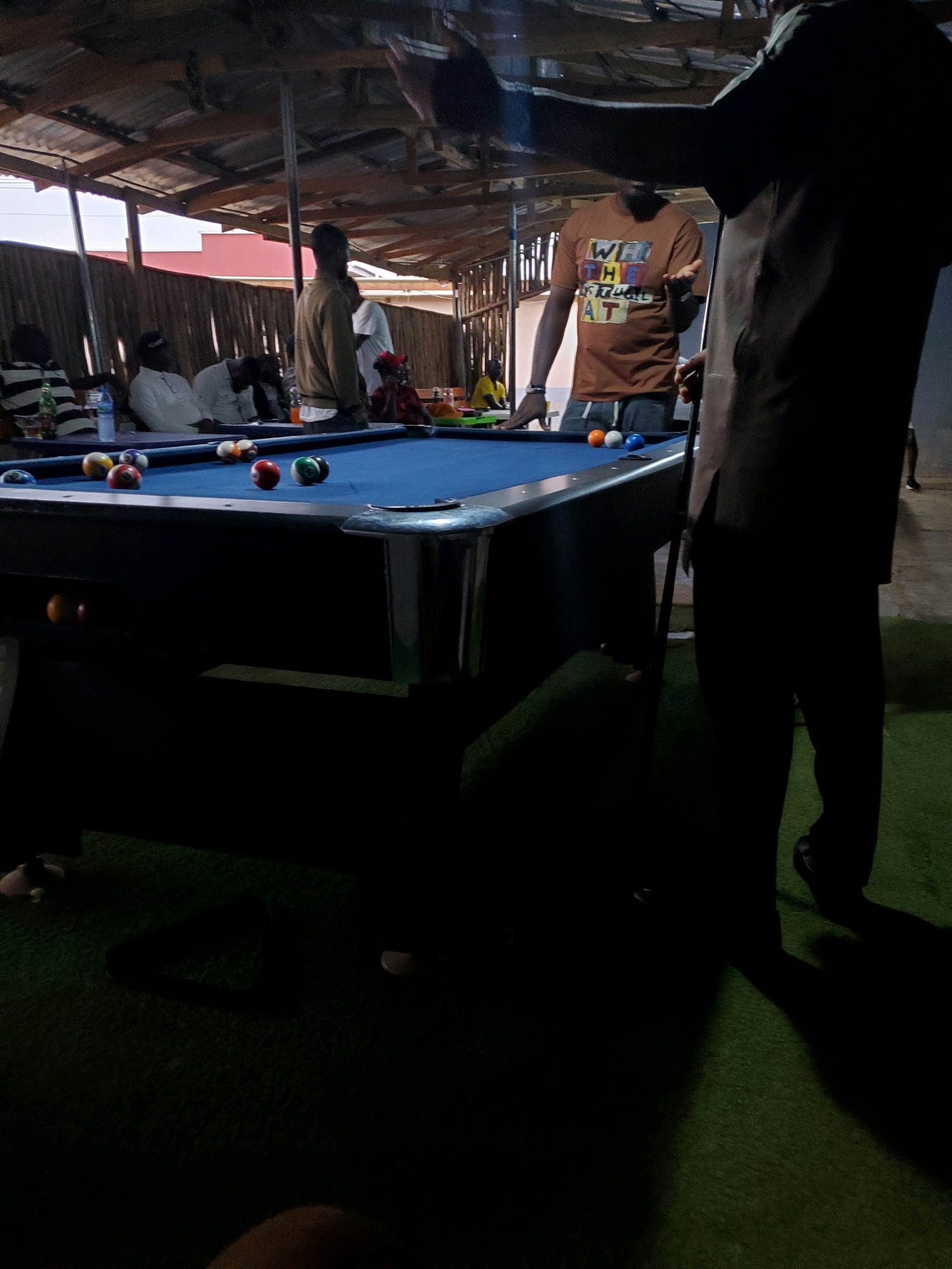 In Jos, Nigeria, a group of individuals are gathered around a pool table situated outdoors under a corrugated metal roof supported by wooden beams. The pool table is covered in blue felt, with various billiard balls scattered across its surface. Three balls, one orange, one white, and one blue, are prominently visible near the center of the table. Two men are actively engaged with the pool table. One man, wearing a brown t-shirt with the text "WEL THE TEAMS AT," is standing on the left side of the table, gesturing with his left hand. Another man, dressed in dark clothing, stands on the right, holding a pool cue. Several other people are seated in the background, some appearing to be watching the game. The environment suggests a casual gathering place, possibly a bar or recreational spot, with artificial turf on the ground. The lighting indicates it is likely evening or nighttime, with shadows cast across the scene.