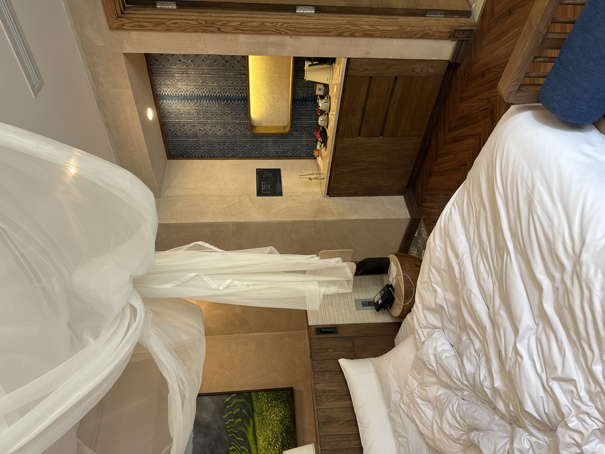 An interior view of a hotel room in Púng Luông, Vietnam, showcases a blend of natural textures and modern amenities. In the foreground, a large bed is made with white linens and pillows, its duvet appearing rumpled. Above the bed, a sheer white canopy or mosquito net is gracefully draped from the ceiling. To the left of the bed, a wooden headboard is visible, above which hangs a framed painting depicting vibrant green and yellow terraced fields. A small, round wooden nightstand to the right of the bed holds a black corded telephone, a black cylindrical object, and a white piece of paper. The walls surrounding the bed area are textured in an earthy brown-tan hue.
To the right, a dedicated mini-bar or coffee station is built into the wall. It features light-colored stone countertops over dark wooden cabinets with drawers and doors. The back wall of this station is adorned with a dark blue patterned wallpaper or fabric, displaying geometric designs. Mounted above the counter is a horizontal, rectangular light fixture emitting a warm yellow glow. On the countertop, various items are arranged: a glass water bottle, multiple pieces of fruit (including dragon fruit and a small orange), two white cups and saucers, a black tray, and a white electric kettle with a coffeemaker. A small black information plaque is affixed to the wall to the left of this station. The floor throughout the visible area is dark wood, laid in a herringbone pattern. A wooden door frame is partially visible on the far right, suggesting an adjacent room or entrance. The ceiling is white, with an air vent visible towards the upper right.
