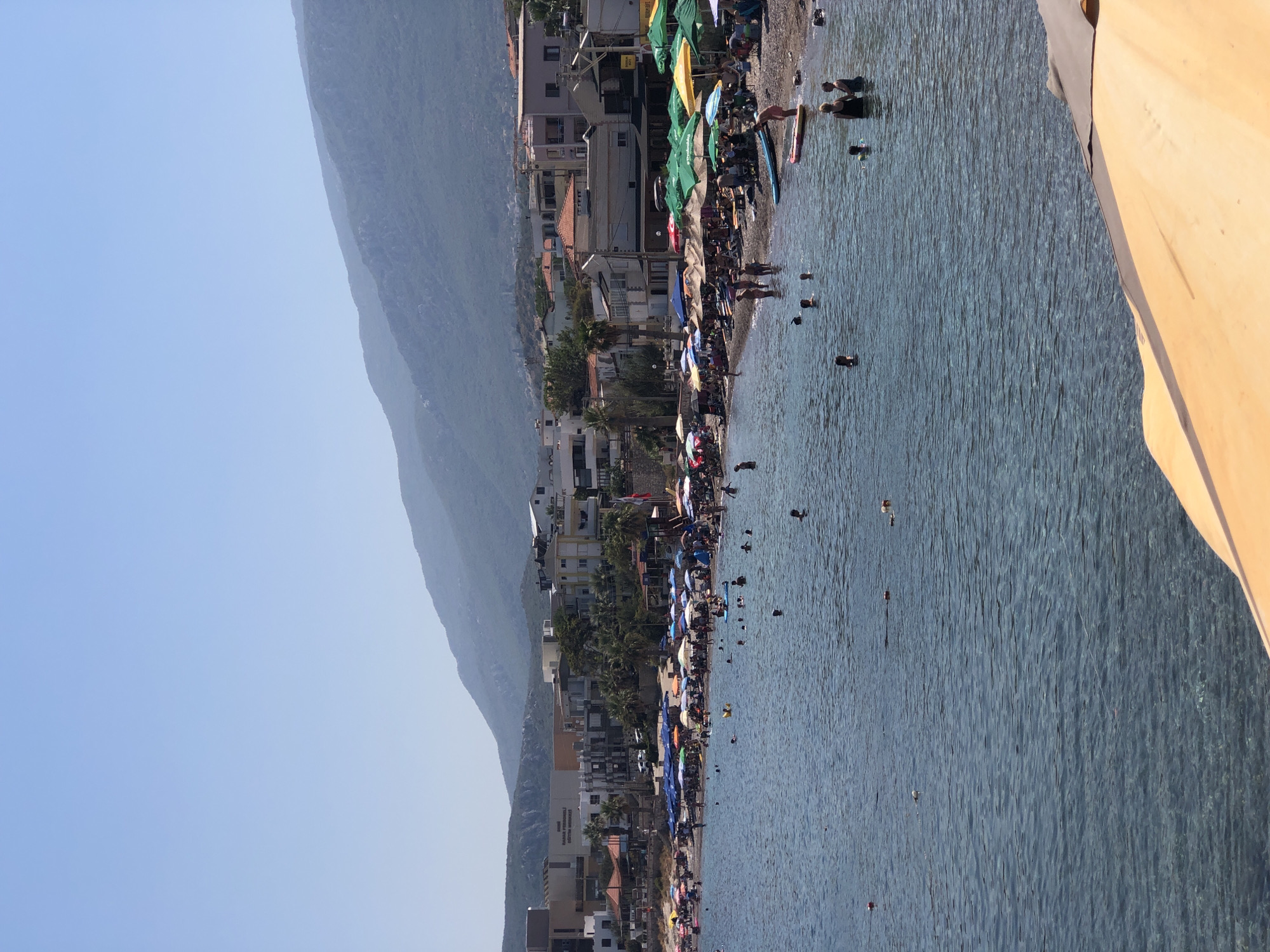 This is a high-angle, eye-level, full, outdoor, vertical, medium shot of a crowded beach and town in Karaburun, Turkey. The water is a clear, shimmering blue, with several people swimming and wading near the shore. The beach is lined with rows of colorful umbrellas and lounge chairs, and a strip of buildings, including hotels and shops, are visible behind the beach. In the background, misty blue mountains rise towards a clear, pale blue sky. The scene appears to be a typical summer day at a popular tourist destination.