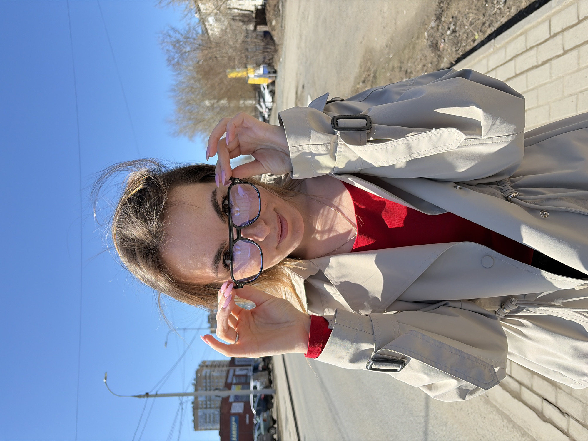 A woman stands outdoors on a sunny day, likely in Izhevsk, Russia. She is wearing a beige trench coat over a red top and has black-framed glasses, which she is adjusting. The background shows a city street with buildings, parked cars, trees without leaves, a street lamp, and a clear blue sky. The surface the woman stands on appears to be a paved sidewalk or road.