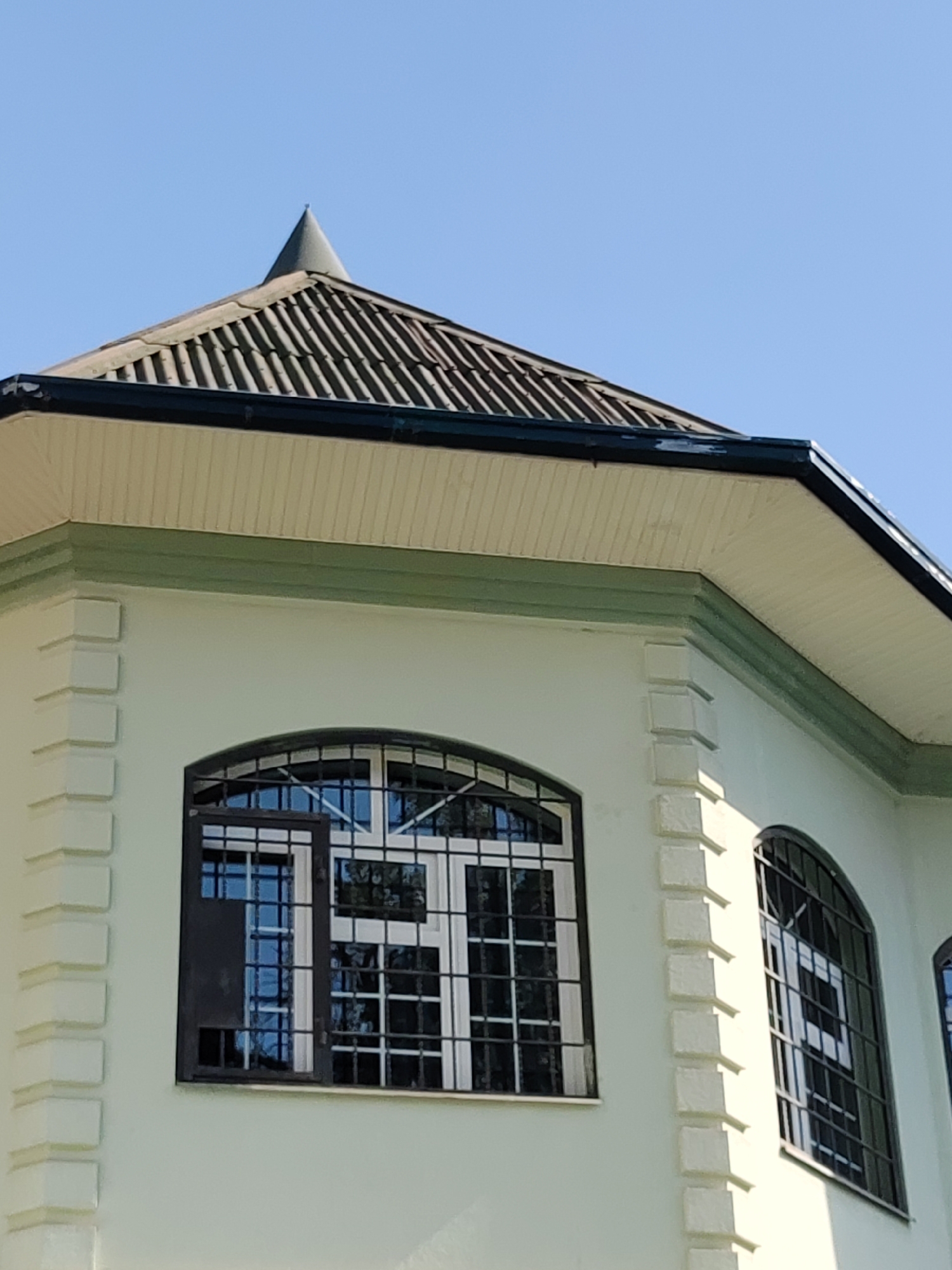The image shows a close-up, low-angle shot of the upper section of a light-green building with white corner detailing. The building features arched windows with black metal bars. The roof is dark gray and tiled, with a pointed spire at its peak. The sky is a clear, bright blue. This building appears to be the Tashkent Television Tower, a prominent landmark in Tashkent, Uzbekistan.