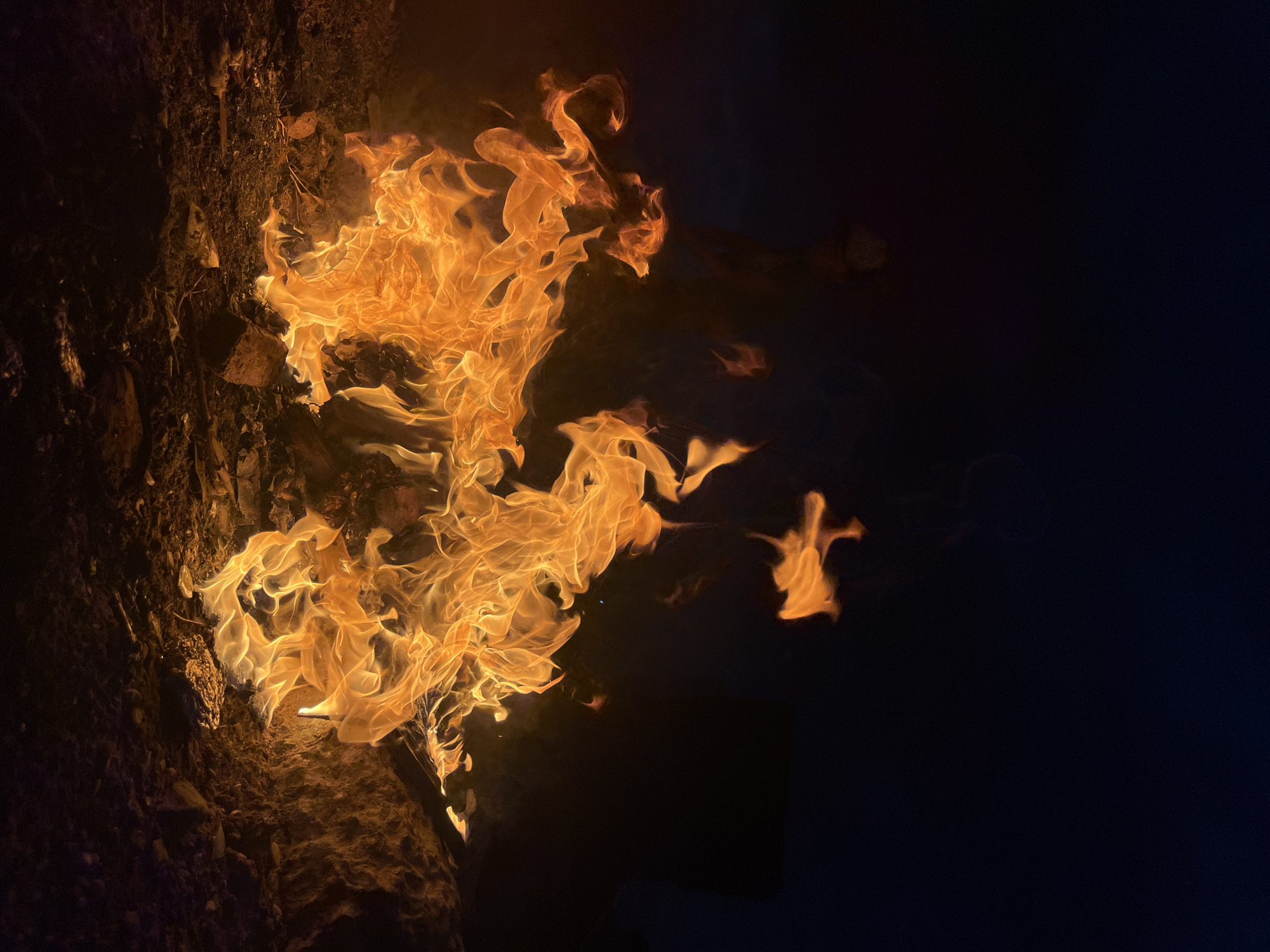 A close-up, low-angle shot captures a campfire burning intensely at night in Kaspiysk, Russia. The flames, predominantly shades of bright orange and yellow, are dynamically shaped by the wind, creating swirling patterns and licking upwards. Wisps of smoke are visible against the dark, inky sky, which takes up the upper portion of the frame. The ground beneath the fire consists of dark earth, scattered rocks, and charred debris. In the background, barely discernible shapes suggest the presence of distant structures or figures, softened by the darkness and the glow of the fire. The overall impression is one of raw, untamed energy emanating from the central blaze.