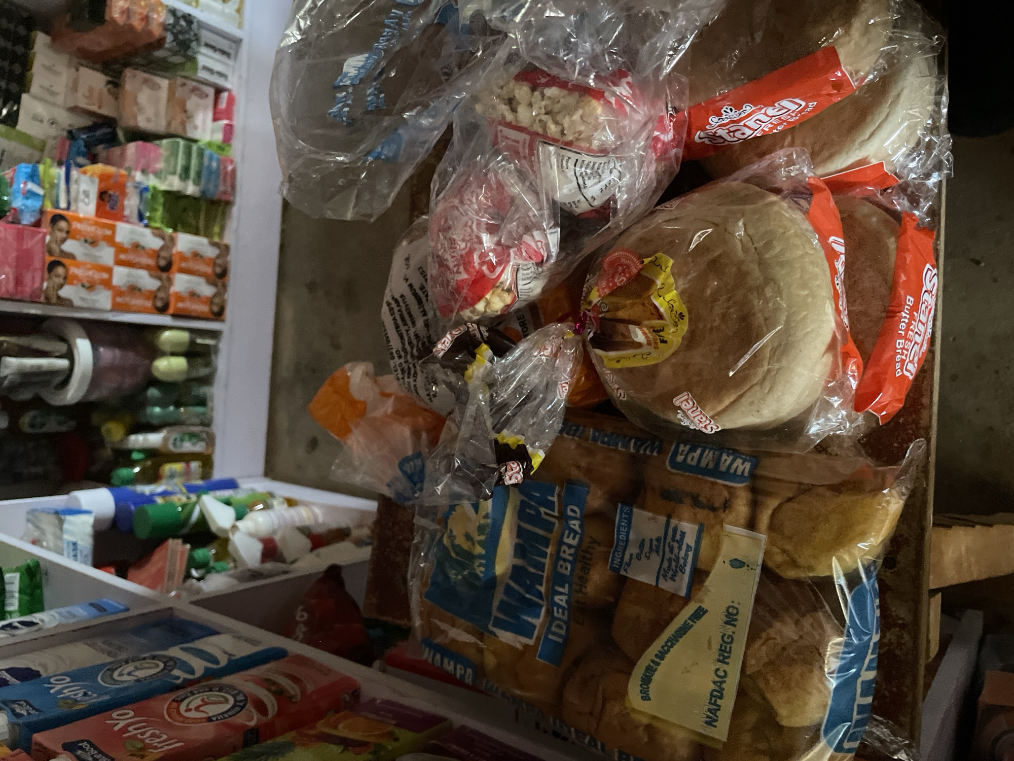 The image shows a display of food items in a Nigerian shop. The foreground features packaged bread, with brands like "Wampa Ideal Bread" and "Stanel Butter Bread" clearly visible. There are also packages of popcorn and other snacks. In the background, shelves are stocked with various goods, including boxes of what appear to be toiletries or personal care items, and bottles of liquid. The overall scene suggests a retail environment, likely a small convenience store or market stall. The lighting is somewhat dim, hinting that it might be indoors or overcast. The presence of NAFDAC registration numbers on the food packaging indicates regulatory compliance within Nigeria. The items are arranged in a somewhat cluttered manner, typical of such retail settings.