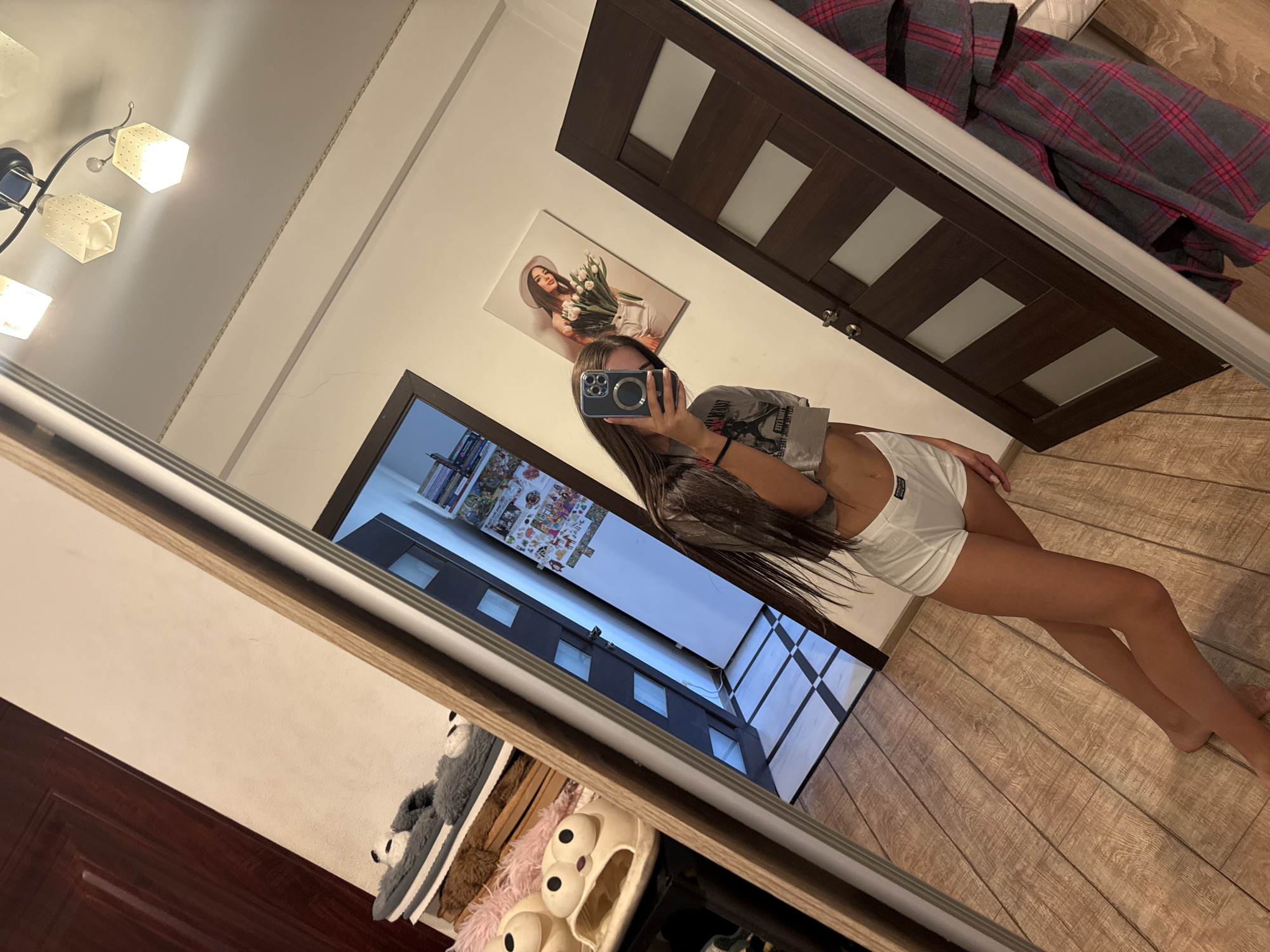 A young woman with long brown hair takes a mirror selfie in a room with light wood-plank flooring. She is wearing a grey crop top and white shorts. She is holding a smartphone in her right hand, which is angled to capture her reflection and a portion of the room. The room features a large mirror, a wooden door with frosted glass panels, and a framed picture on the wall. To the left of the mirror, visible in the reflection, are several pairs of slippers on shelves. A ceiling light fixture with three lamps is visible in the upper portion of the mirror's reflection. The overall impression is of a domestic interior, likely a bedroom or dressing area.