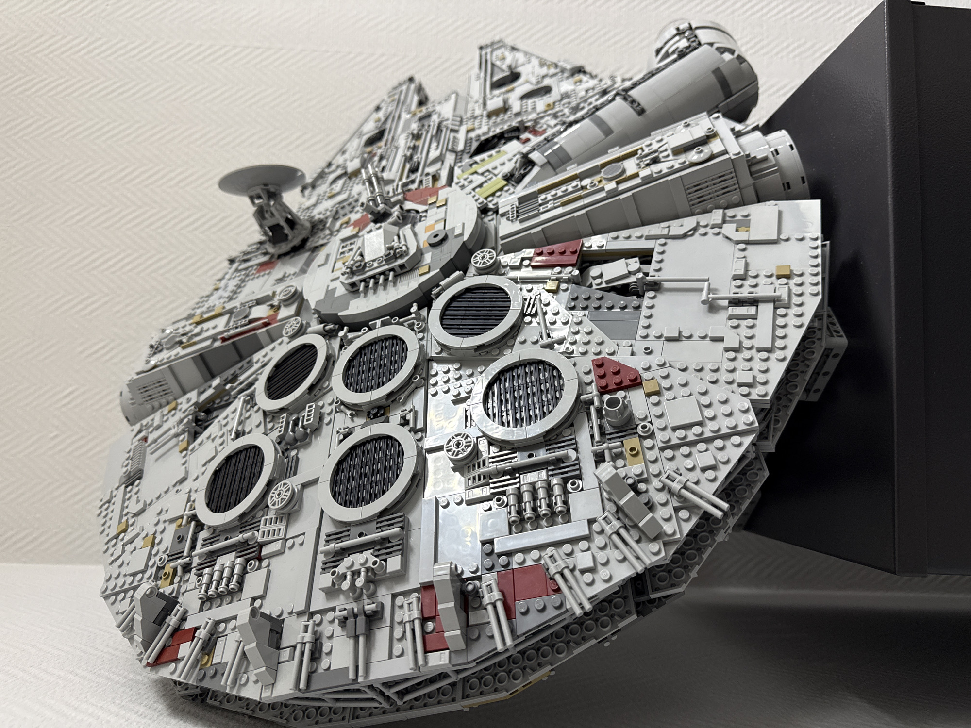 The image shows a detailed Lego model of the Millennium Falcon, seemingly resting on a surface. The model is predominantly gray and off-white, with intricate detailing and various circular elements. It is positioned against a white, subtly textured background. A black, rectangular object or surface is visible on the right side of the image, partially obscured by the model. There is no indication of the location being Krasnoyarsk, Russia, nor any people or actions visible in the scene.