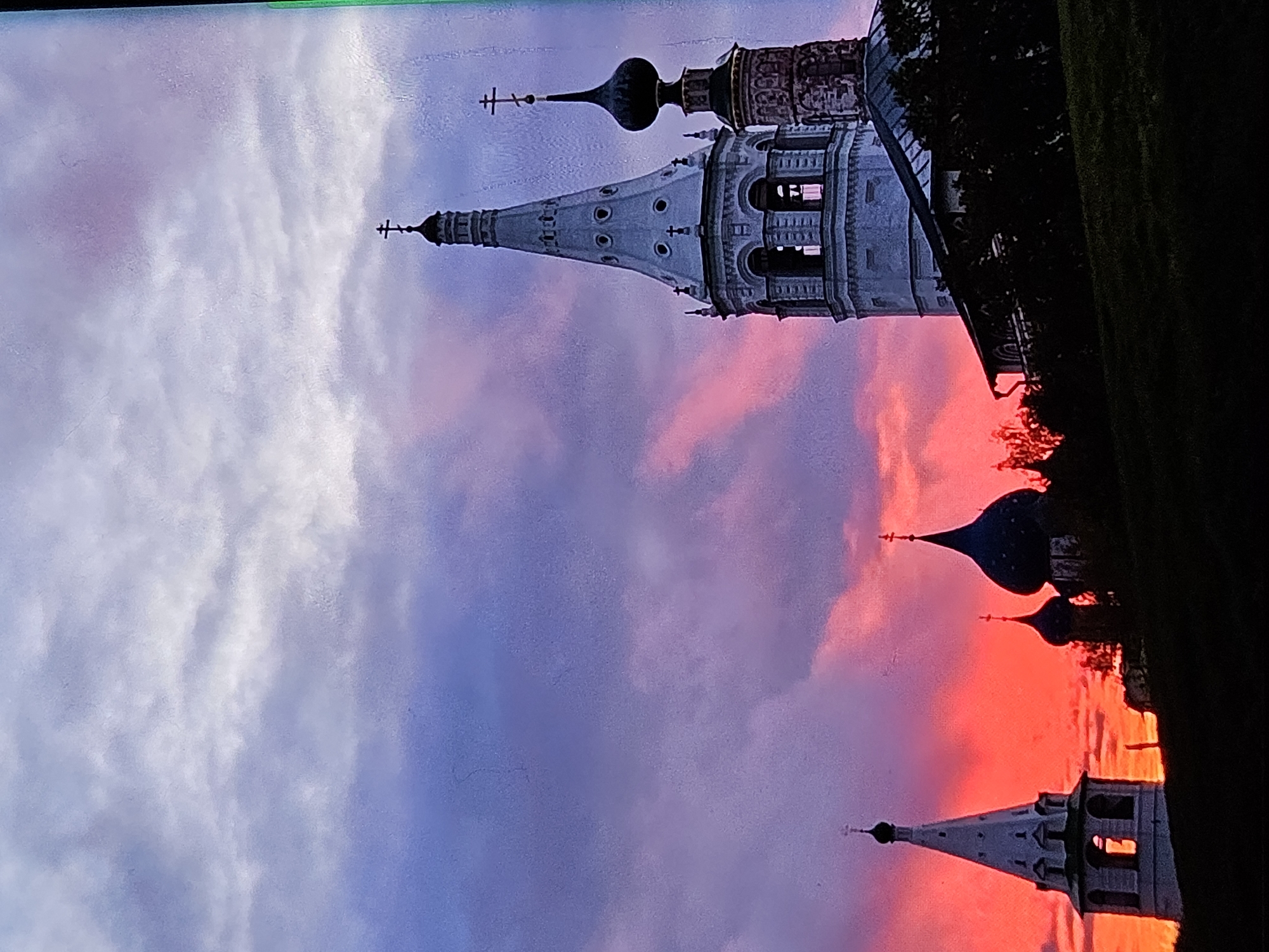 The image shows a silhouette of the Church of St. John the Baptist, part of the Church of St. John the Baptist complex in the city of Vladimir, Russia, against a dramatic sunset sky. The sky is a vibrant display of colors, transitioning from deep purples and blues at the top to fiery oranges and pinks near the horizon. Several church towers, each topped with a cross, are visible against this colorful backdrop. The foreground consists of a dark, silhouetted grassy hill. The scene is peaceful and evokes a sense of natural beauty and architectural grandeur.