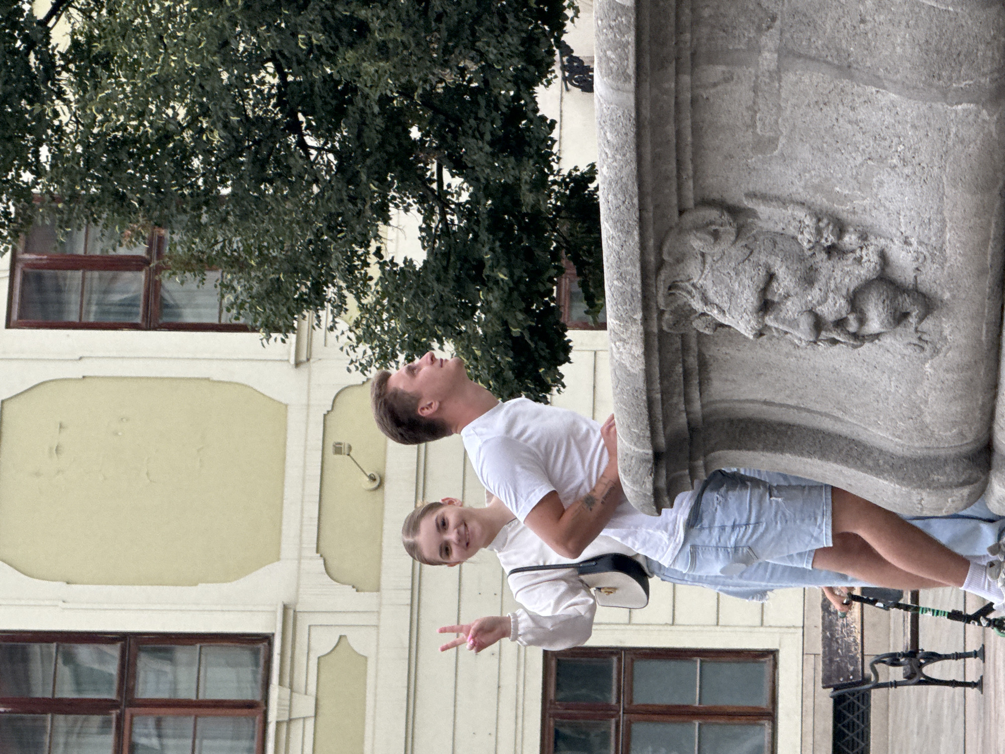 Two young people, a man and a woman, are posing for a photo outdoors. The man is looking up and to his left, with his mouth slightly open. He is wearing a white t-shirt and blue denim shorts. The woman is smiling and giving a peace sign with her left hand. She is wearing a white long-sleeved top and a dark crossbody bag. They are standing in front of a building with light yellow walls and dark-framed windows. Behind them, to the left, is a large tree with dark green foliage. To the right, there is a large stone structure, possibly a fountain or monument, with a carved lion's head visible. The scene appears to be set in a city, likely in Bratislava, Slovakia, given the architectural style of the building and the presence of the stone monument. The lighting suggests it is daytime, possibly overcast or with soft sunlight, as there are no harsh shadows. The overall mood seems cheerful and relaxed, with the woman's smile and peace sign indicating a positive interaction. There is no visible text in the image.