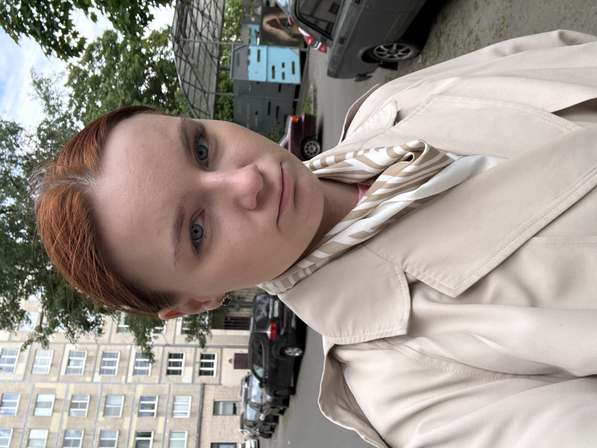 A person with short reddish-brown hair, light skin, and blue eyes is taking a selfie outdoors. The person is wearing a light beige trench coat and a beige and white patterned scarf. Behind the person, there is an apartment building with white-framed windows and a row of parked cars. A gray car is also visible in the upper right corner of the frame. Based on the architectural style of the apartment building, it could be located in Saint Petersburg, Russia. The scene appears to be set on a street.
