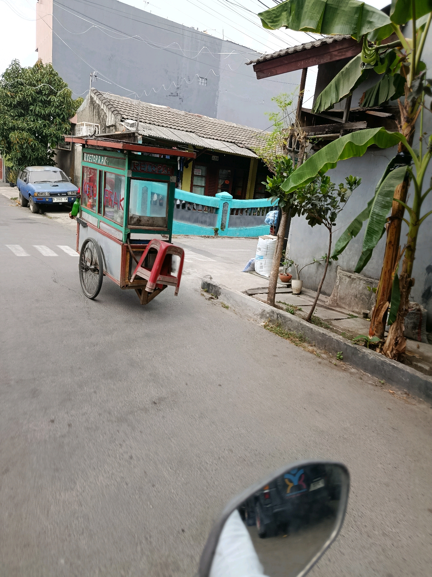 The image captures a street scene in Bekasi, Indonesia, featuring a food cart and its surroundings. In the foreground, a mobile food cart, primarily green and red, is parked on the side of an asphalt road. The cart has wheels and a canopy, with visible text on its side that appears to read "KETOPRAK". A red plastic stool is attached to the cart.
To the left of the cart, a blue car is parked on the roadside. Behind the car and the cart, there are buildings and vegetation. A large, leafy green tree stands prominently to the left of the scene. The buildings have tiled roofs and a mix of gray and pastel-colored walls. A distinctive turquoise-colored fence or railing runs along the front of one of the buildings. In the background, a tall, modern building with a gray facade is visible.
The image is taken from the perspective of someone on a motorcycle, as indicated by the reflection in the rearview mirror visible in the bottom right corner. The mirror reflects a blurred image of the rider and a portion of the motorcycle. The overall environment suggests a typical urban street in Indonesia, with residential buildings and food vendors.