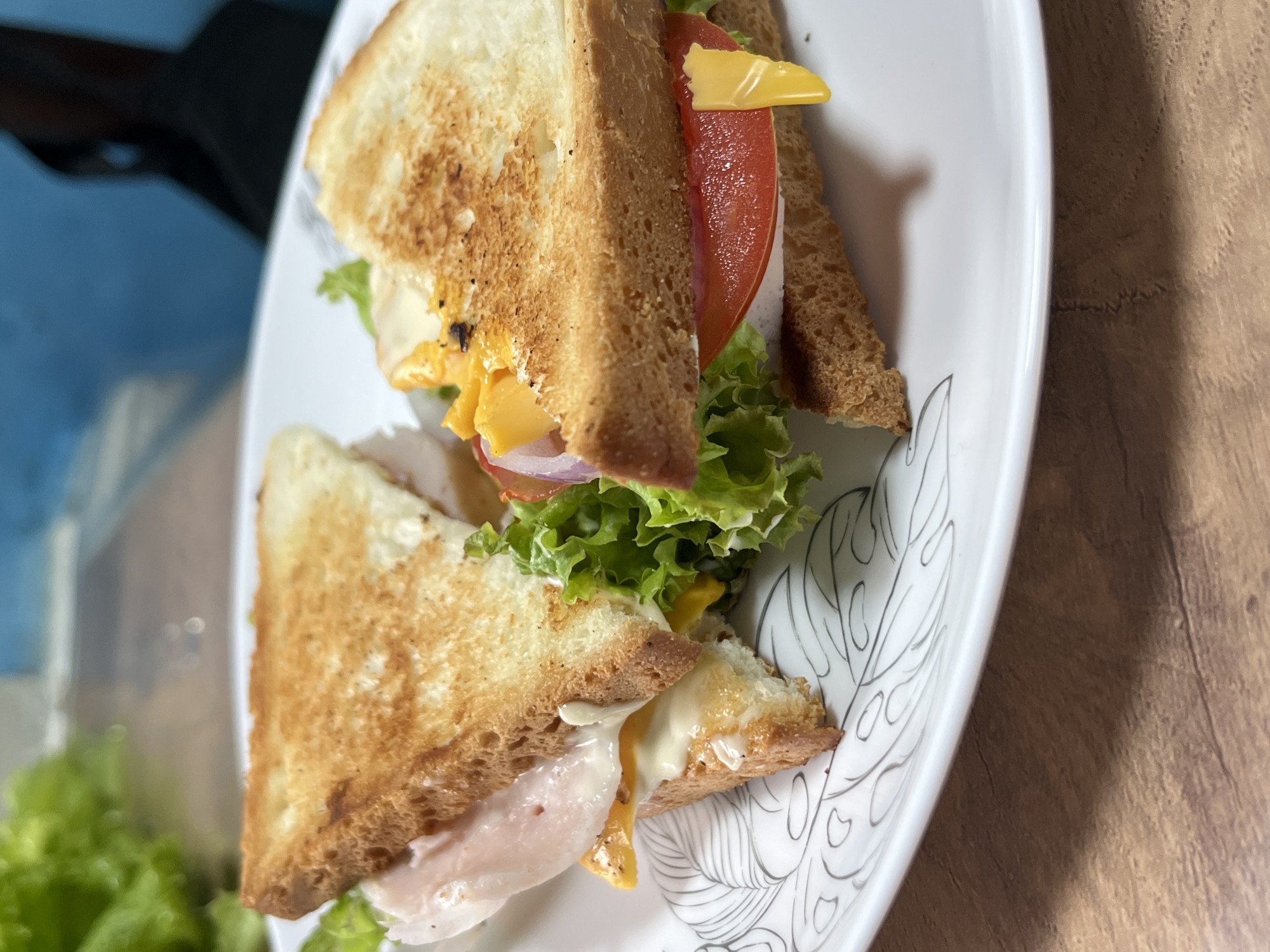 A close-up, eye-level shot showcases two halves of a sandwich on a white plate with a black leaf pattern. The sandwiches are made with toasted bread and filled with lettuce, tomato, cheese, sliced red onion, and what appears to be deli meat and mayonnaise. The plate sits on a light brown wooden table. In the blurred background, a bright green head of lettuce is visible. There is no visible information in the image to suggest it is located in Nizhny Tagil, Russia.