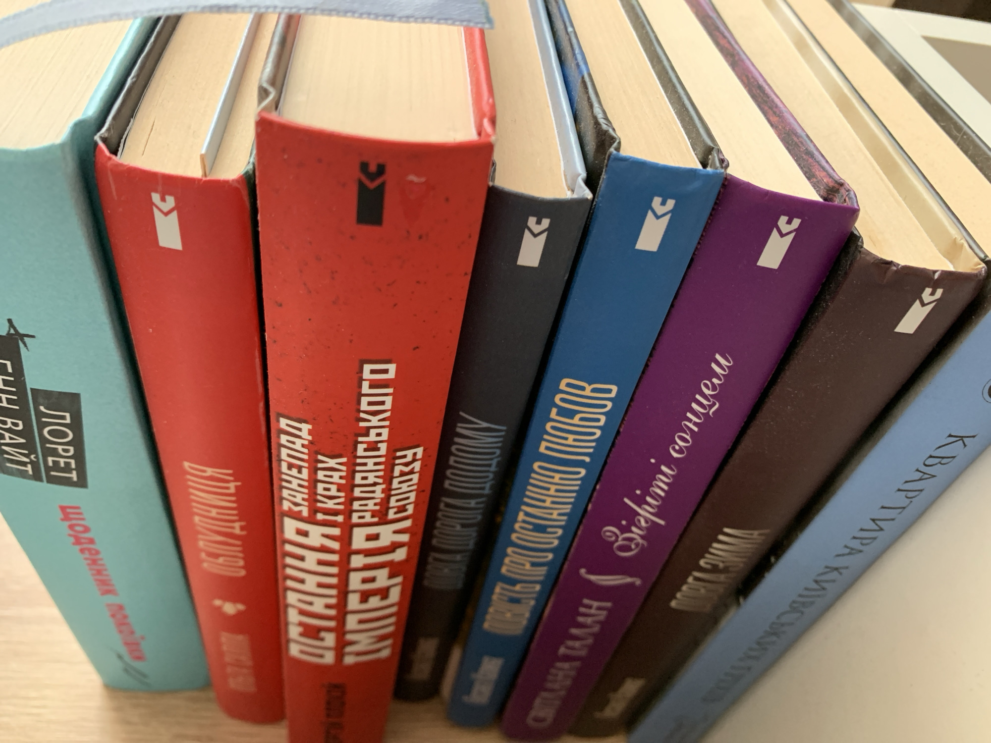 The image shows a stack of books. The book titles are in Ukrainian. The top book is light blue and has the title "Дорет Байт" on it. The second book from the top is red and has the title "Облудниця" on it. The third book from the top is also red and has the title "ОСТАННЯ І КРАХ ІМПЕРІЯ РАДЯНСЬКОГО СОЮЗУ" on it. The fourth book is dark blue and has the title "ДОРІГА ДО ДОМУ" on it. The fifth book is a brighter blue and has the title "ПОКІСТЬ ПРО ОСТАННЮ ЛЮБОВ" on it. The sixth book is purple and has the title "СВІТЛА ТАЛАН" on it. The seventh book is dark brown and has the title "ПОРГА ЗИМА" on it. The bottom book is light blue and has the title "КВАРТИРА КНИЖКОВИХ МІСТ" on it. There is no information available to confirm the location in Ternopil, Ukraine.