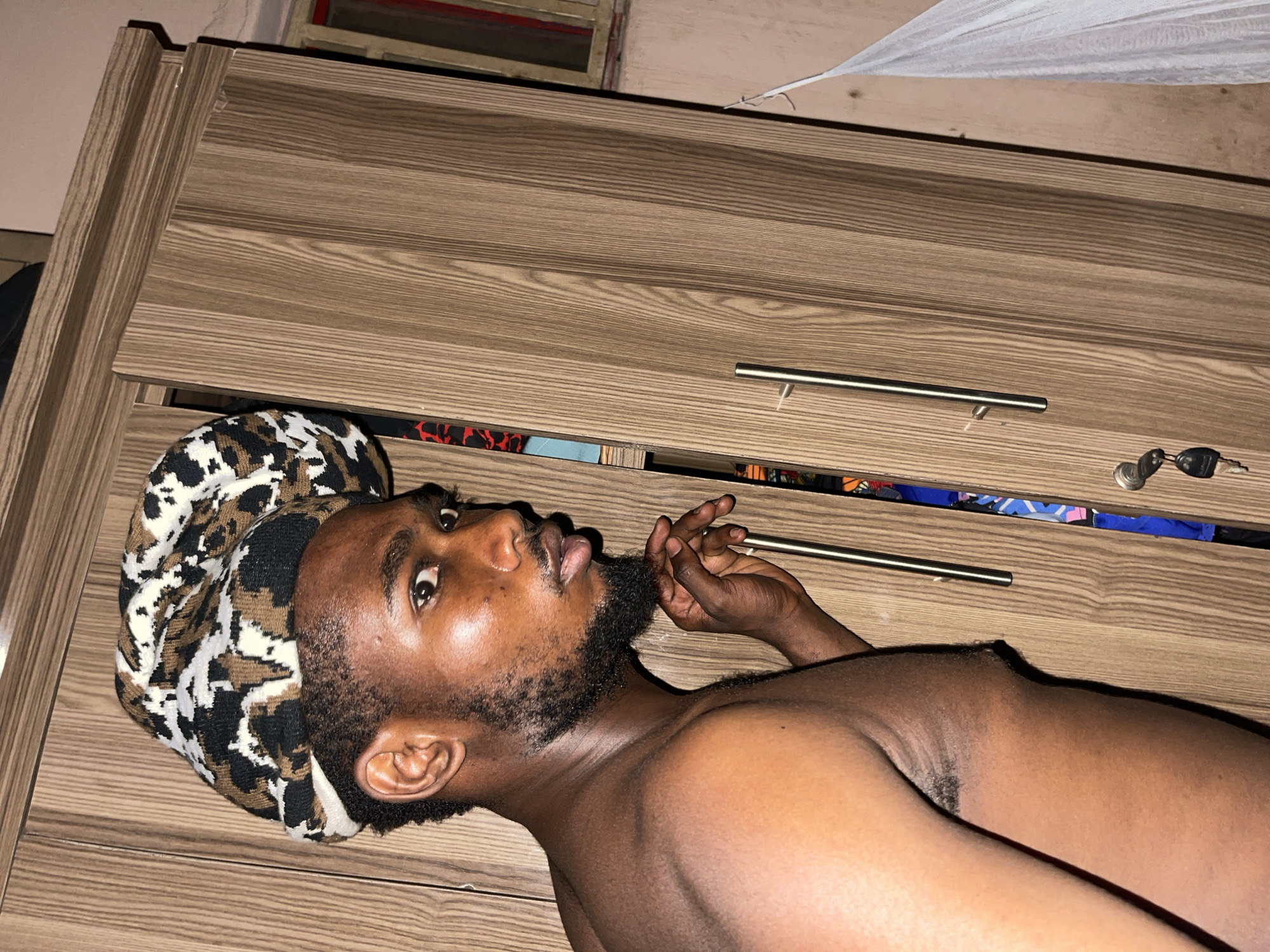 A person with dark skin and a beard is lying on what appears to be a wooden dresser or cabinet. The person is wearing a printed head covering. A set of keys is visible on the right side of the dresser. The location is not evident from the image. There is no information about the city of Muchiya.