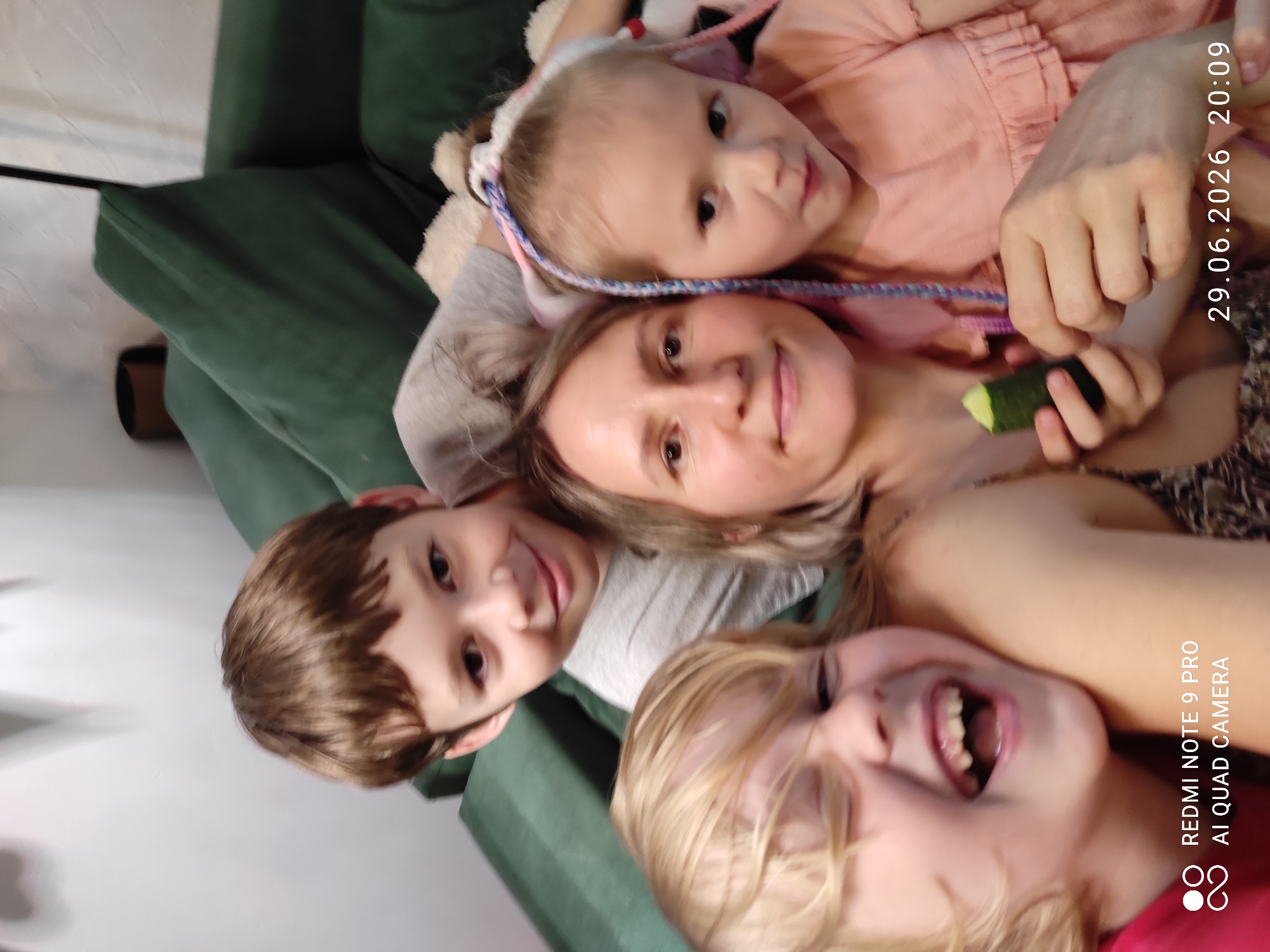 This is a close-up, overhead shot of a mother and three children on a green couch. The mother is lying down, with a baby resting on her chest. To her left is a young boy, and to her right is another child with blonde hair. The children appear to be laughing and interacting playfully. The scene suggests a casual, indoor family moment.
The baby, wearing a headband with braided strings, looks directly at the camera. The mother has a gentle smile. The boy on the left is also smiling broadly, and the child on the right has their mouth open in laughter.
The image is taken from a high angle, looking down at the subjects. The lighting suggests it is daytime, possibly indoors.
Visible text on the right side of the image reads: "29.06.2026 20:09" and "REDMI NOTE 9 PRO AI QUAD CAMERA".