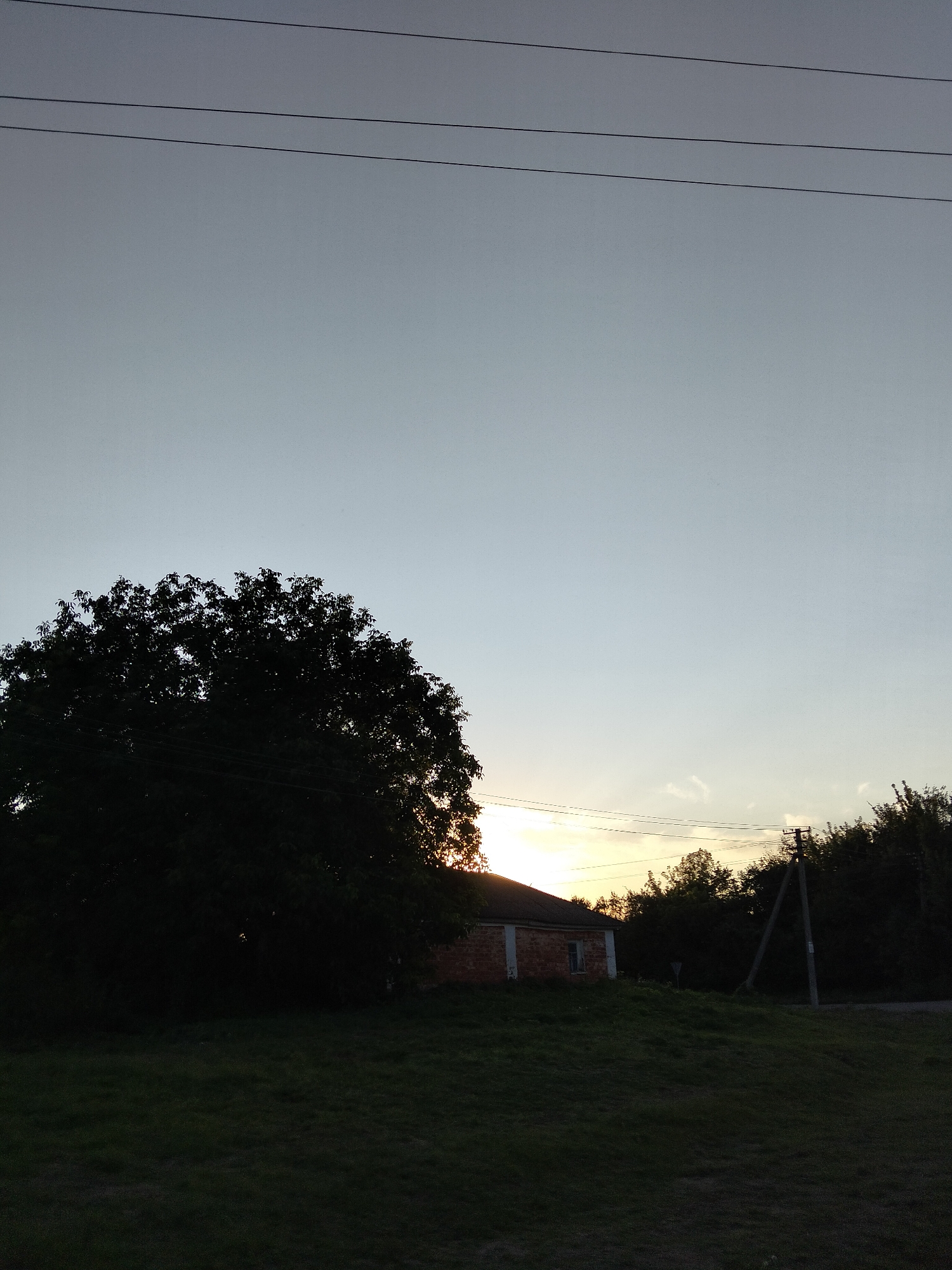 The image depicts a rural scene at dusk in Kliushnykivka, Ukraine. In the foreground, a dark, grassy field slopes upwards. To the left, a large, silhouetted tree with dense foliage dominates the mid-ground. Behind the tree and slightly to the right, a small, brick house with white trim and a single window is visible. The house's roof is visible, and the setting sun is peeking from behind the trees to the right of the house, casting a warm glow on the horizon. Power lines run horizontally across the upper portion of the image. Further to the right, a tall, slender utility pole with a triangular structure at its base stands against the twilight sky. The background consists of a line of dark trees. The sky is a pale gradient of grey to light blue. There are no people visible in the image.