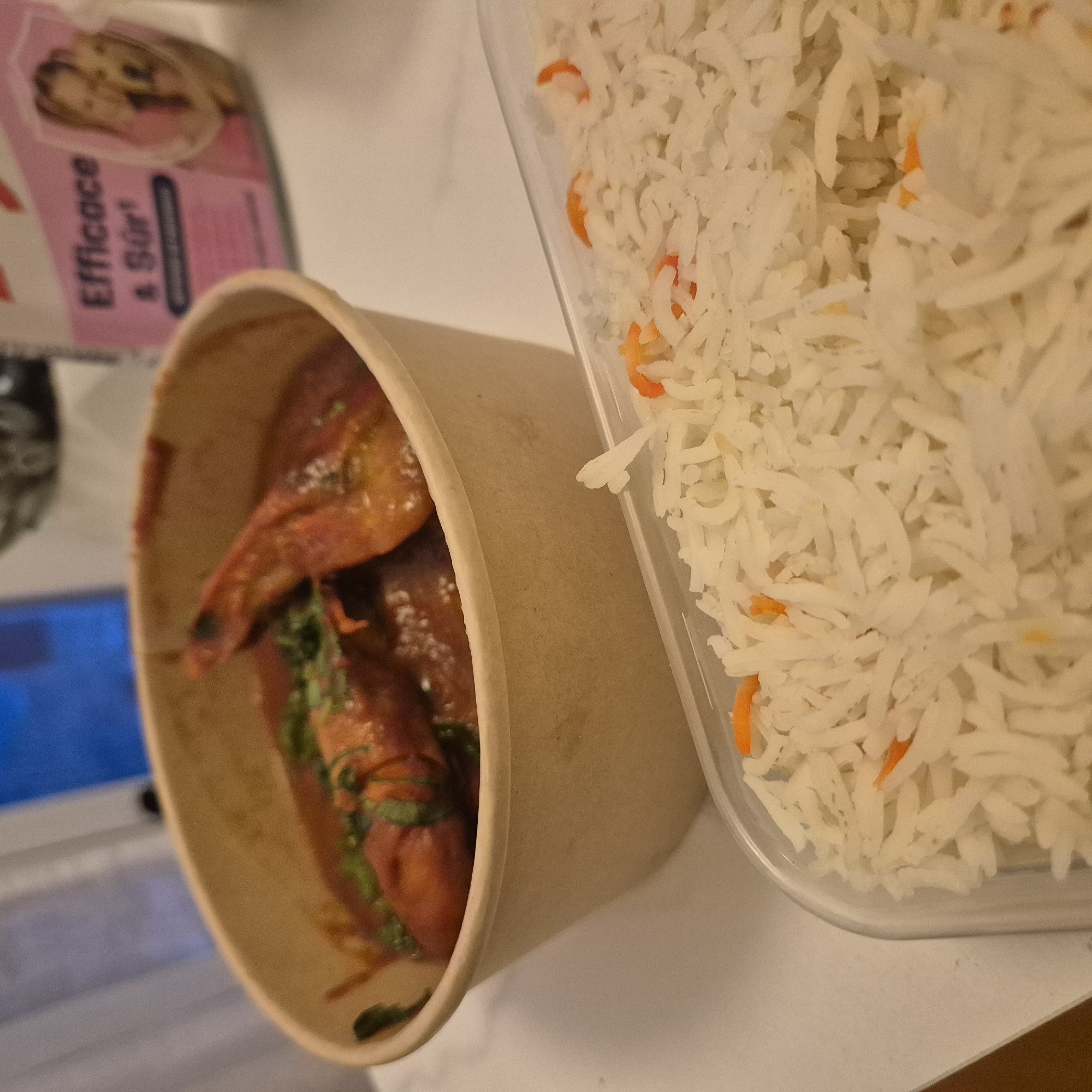 The image shows a takeout meal consisting of rice and curry, likely photographed in Strasbourg, France, based on the packaging visible in the background ("Efficace & Sûr"). The rice, in a plastic container, is predominantly white with occasional orange flecks, possibly carrots. The curry is in a cardboard container and contains what appears to be chicken or meat, accompanied by a red-brown sauce and green herbs. The meal is likely on a table or countertop.