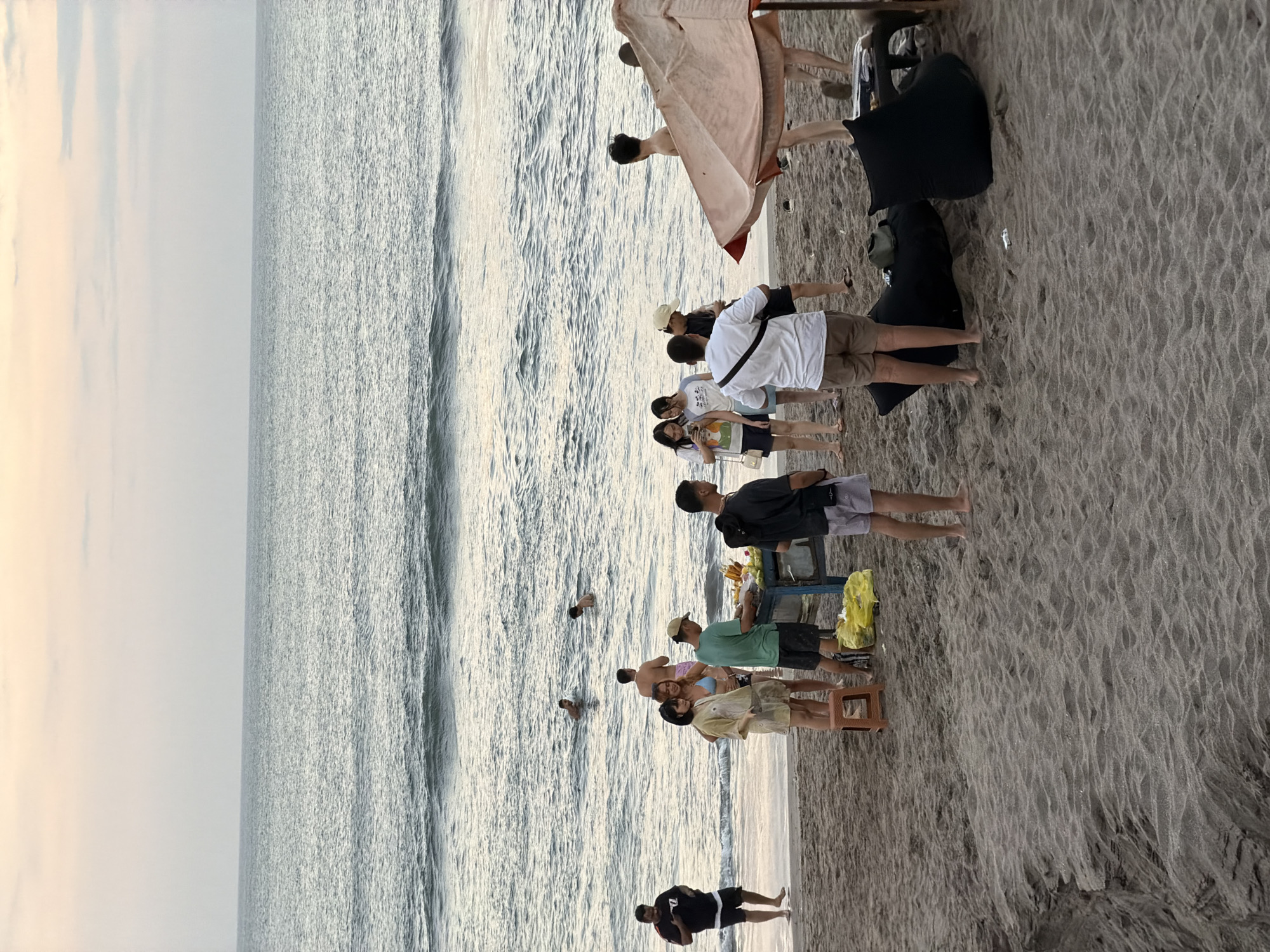 The image depicts a beach scene in North Kuta, Indonesia. Several people are present, some wading in the shallow water and others standing on the sandy shore. A group of individuals are gathered near a cart, possibly a vendor, which is laden with fruits, including bananas. One person is seen using a phone. The water is calm with gentle waves. The sky appears overcast, suggesting either early morning or late afternoon. The overall atmosphere is casual and relaxed, with people enjoying a day at the beach. There is no visible text in the image.