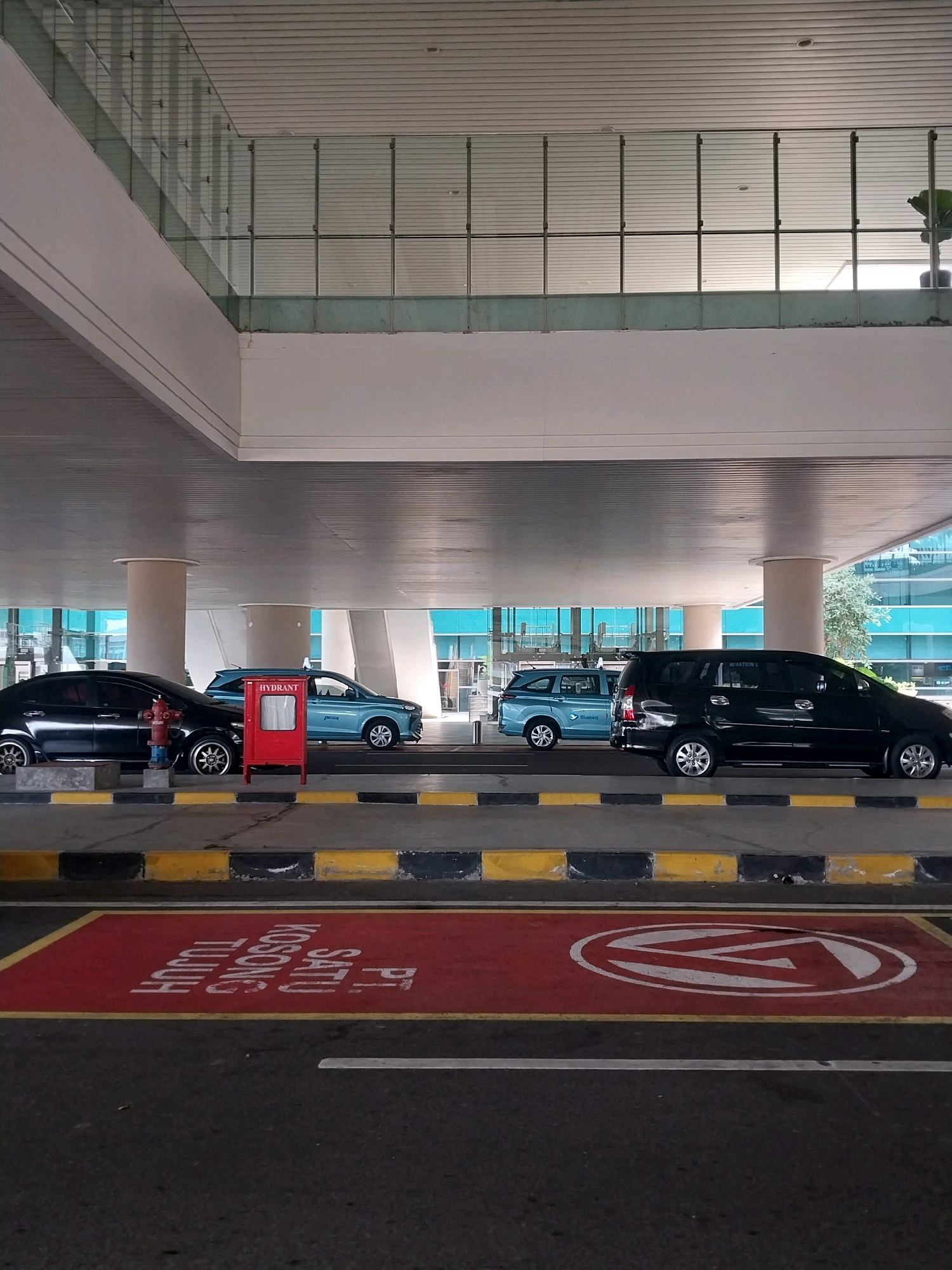 The image depicts an outdoor parking area under a covered structure, likely an airport or a large building, in Palihan, Indonesia. The scene is dominated by concrete pillars supporting the overhead structure and the building's facade, which features extensive glass windows and a white, slatted ceiling. In the foreground, a red zone is marked on the asphalt with yellow and black bordered curbs. Text in white lettering on the red zone reads "PT. 1 SATU KOSONG TUJUH". A white directional arrow is also painted on a separate red zone.
Parked vehicles are visible in the mid-ground. On the left, a black sedan is parked next to a red hydrant. To its right, a light blue hatchback is parked. Further to the right, a dark blue minivan with "Bluebird" branding is visible, and to its right, a black SUV is parked. The environment appears to be well-maintained, with clean asphalt and clear demarcation of parking spaces. The lighting suggests it is daytime, possibly overcast or in shade due to the covered structure. No people are visible in the frame.
