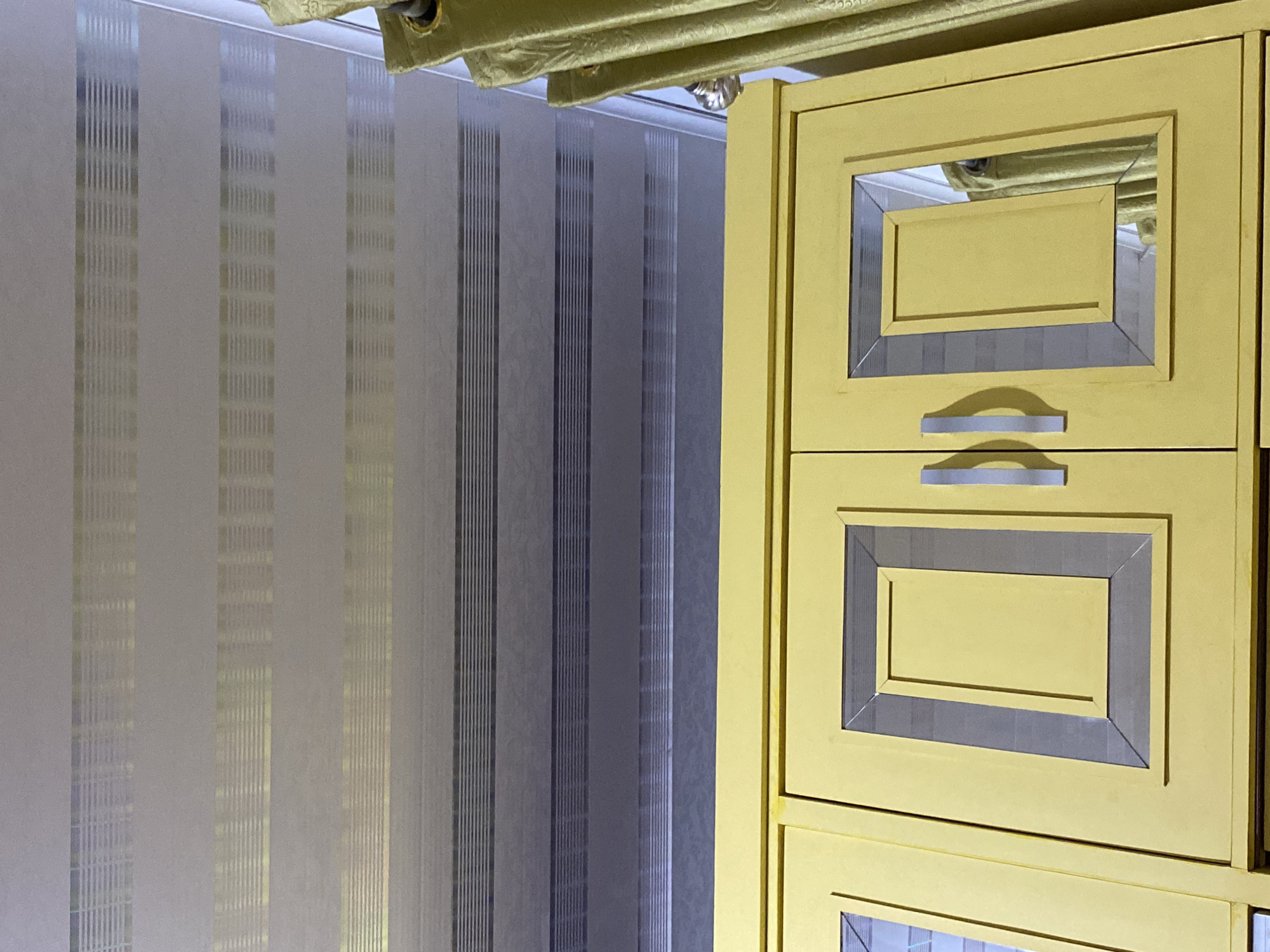 The image shows the interior of a room. In the foreground, the top portion of a bright yellow cabinet with two doors is visible. Each door has a recessed rectangular panel with a silver metallic border. The cabinet has two silver handles. Above the cabinet is a ceiling with horizontal textured panels in alternating shades of gray and off-white with metallic horizontal lines. To the right of the cabinet and ceiling, a portion of a light green curtain is visible. The scene appears to be indoors, likely a domestic setting. No people are present. The location is specified as Lambaro Kueh, Indonesia, though there are no visual cues in the image to confirm this specific location.