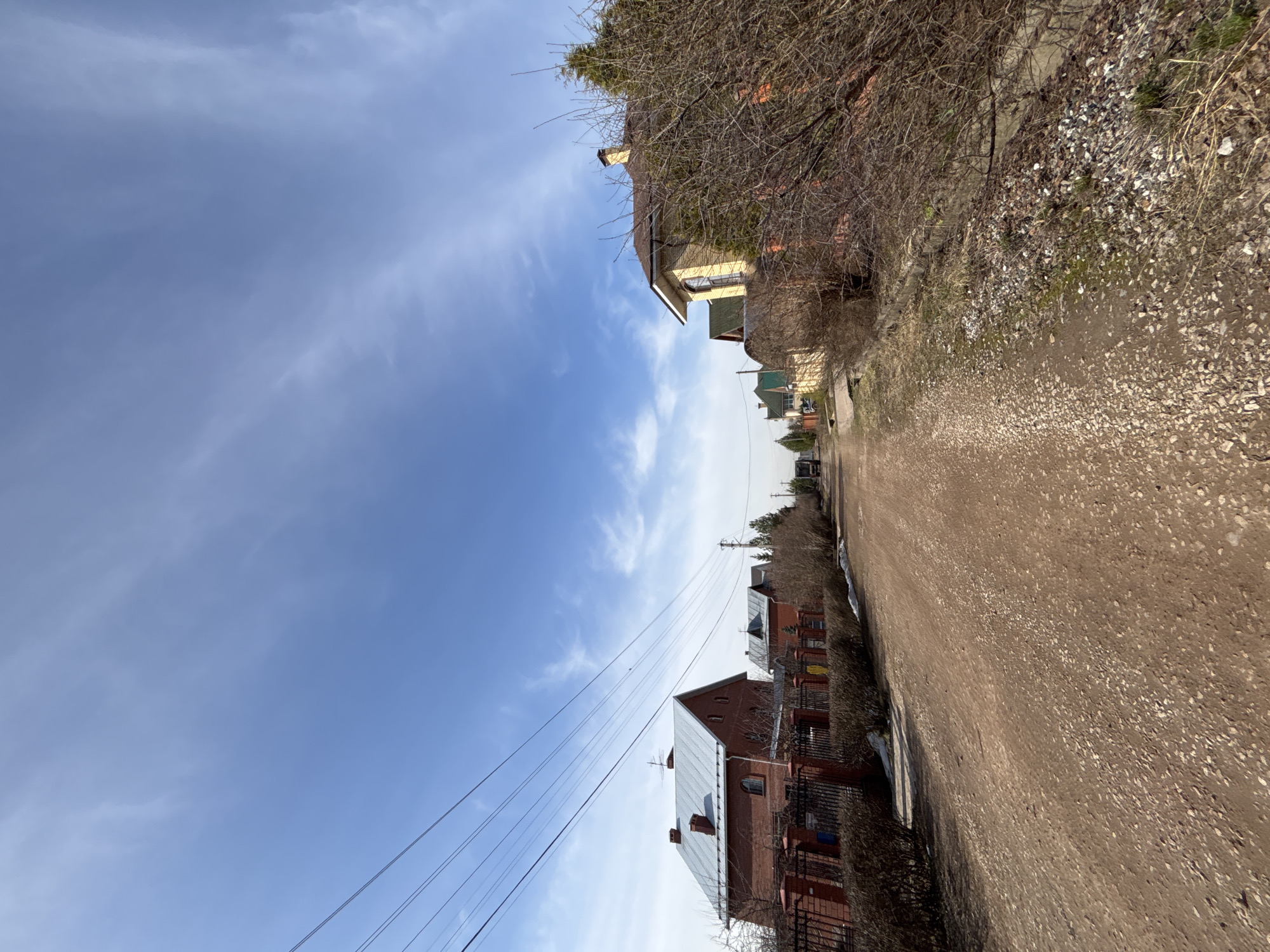 The image depicts a street in the settlement of Ferme, Russia. The view is along a dirt road lined with houses on both sides. The houses on the right are mostly two-story brick buildings with metal roofs, some with fences. The buildings on the left are more varied in style, including wooden structures. Overhead power lines run along the street. The sky is blue with scattered clouds. The foreground is a dirt road, which is uneven. Trees and foliage are visible along the roadside.
