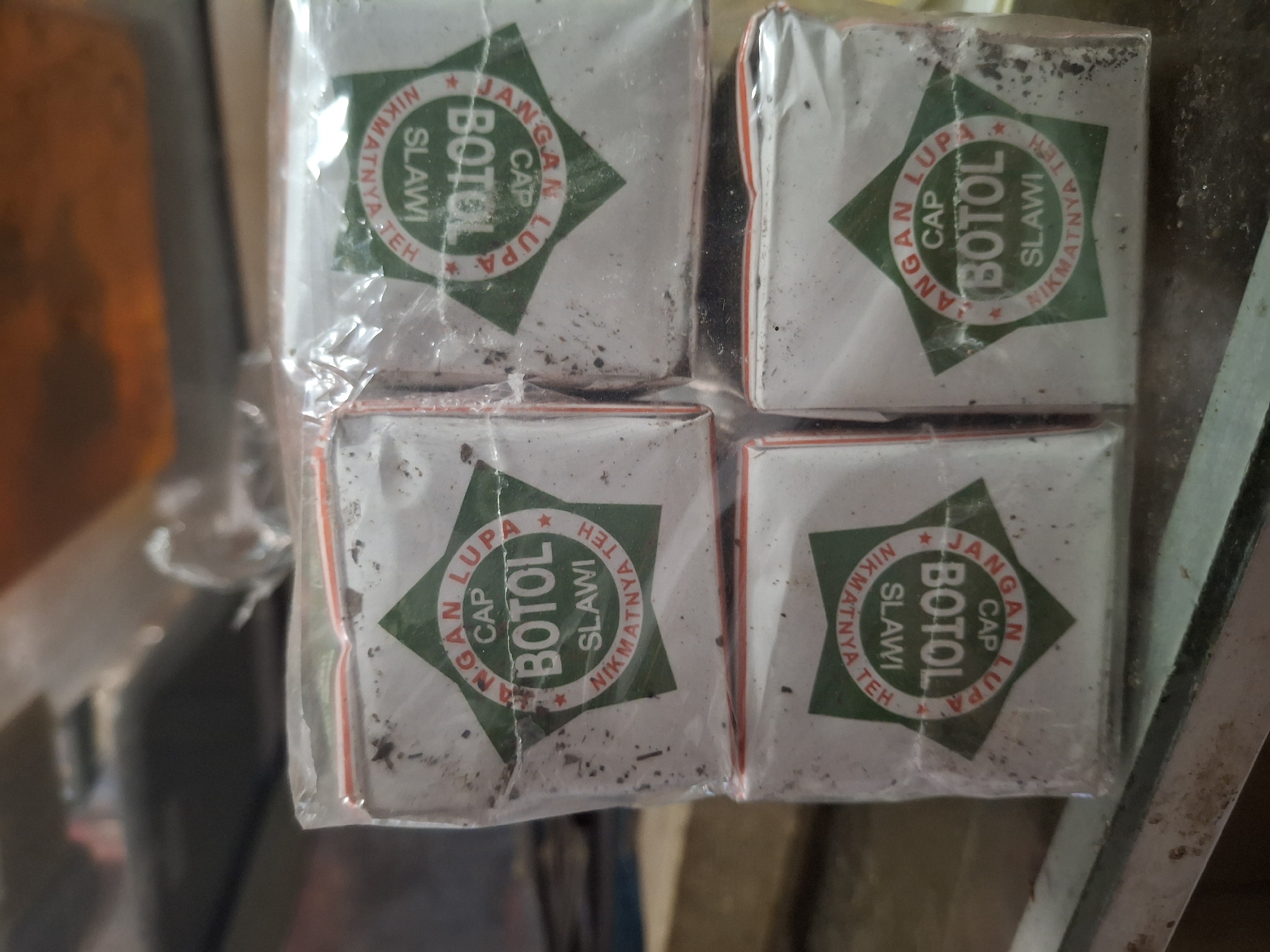 Four individually wrapped boxes of tea are stacked in a 2x2 formation, partially obscuring each other. Each box is white with a green star logo. Inside the star, text reads "CAP BOTOL SLAWI NIKMATNYA TEH DANGAN LUPA". The boxes appear to be dusty and slightly worn, with some visible dirt or debris on the outer plastic wrapping. The background is blurred and appears to be a shelf or counter with indistinct objects. The scene is indoors, and the lighting is moderately bright. The image quality is slightly grainy. This image is not in Biak Kota, Indonesia.
